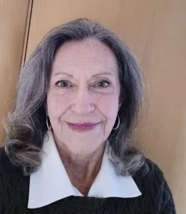Close-up portrait of a smiling middle-aged woman with gray hair, wearing a black top over a white collared shirt, standing in front of a wooden door.