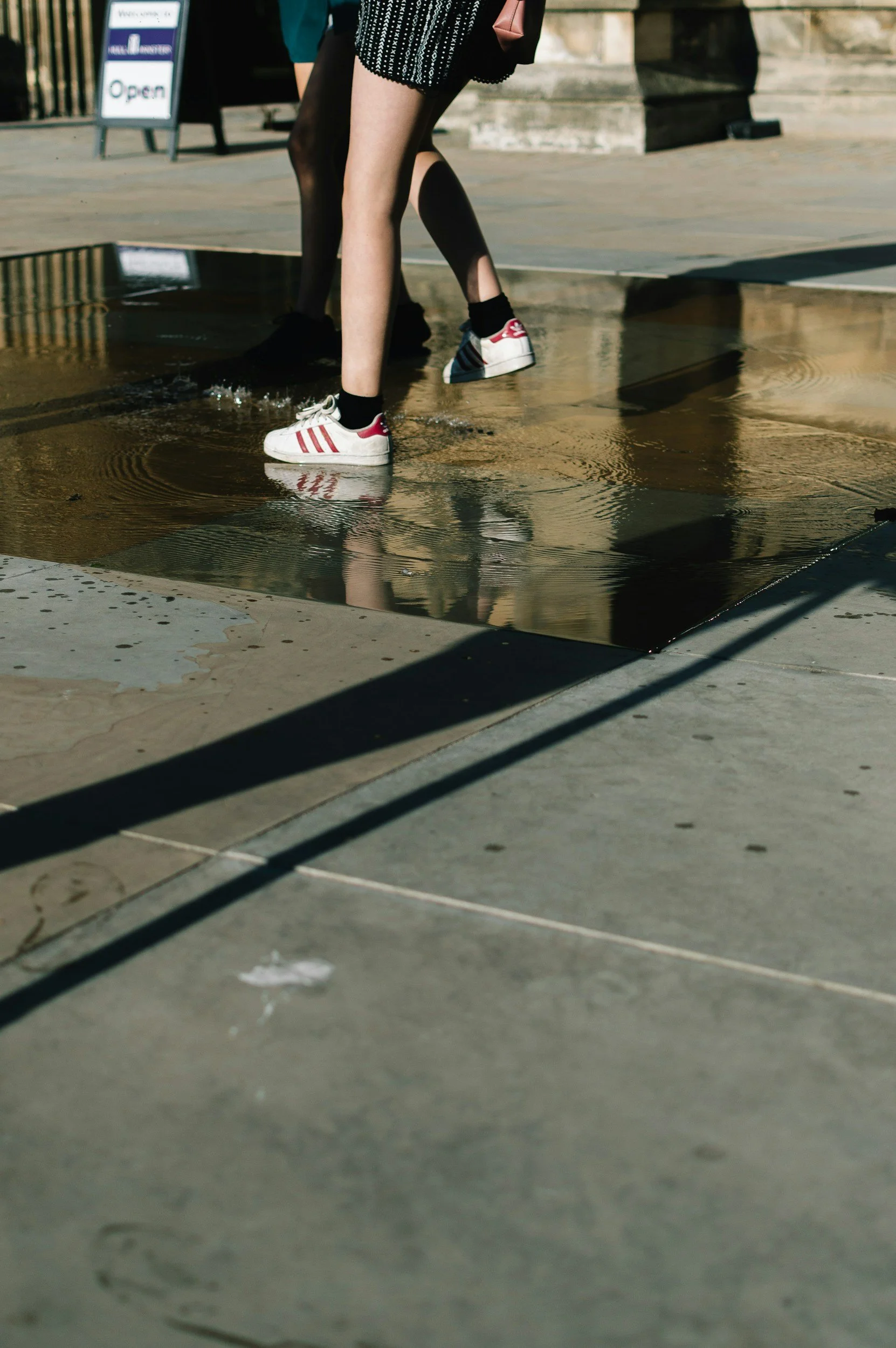 A child wearing white sneakers with red stripes and black socks is stepping over a small puddle on a sidewalk. The shadow of a metal railing is cast on the ground.