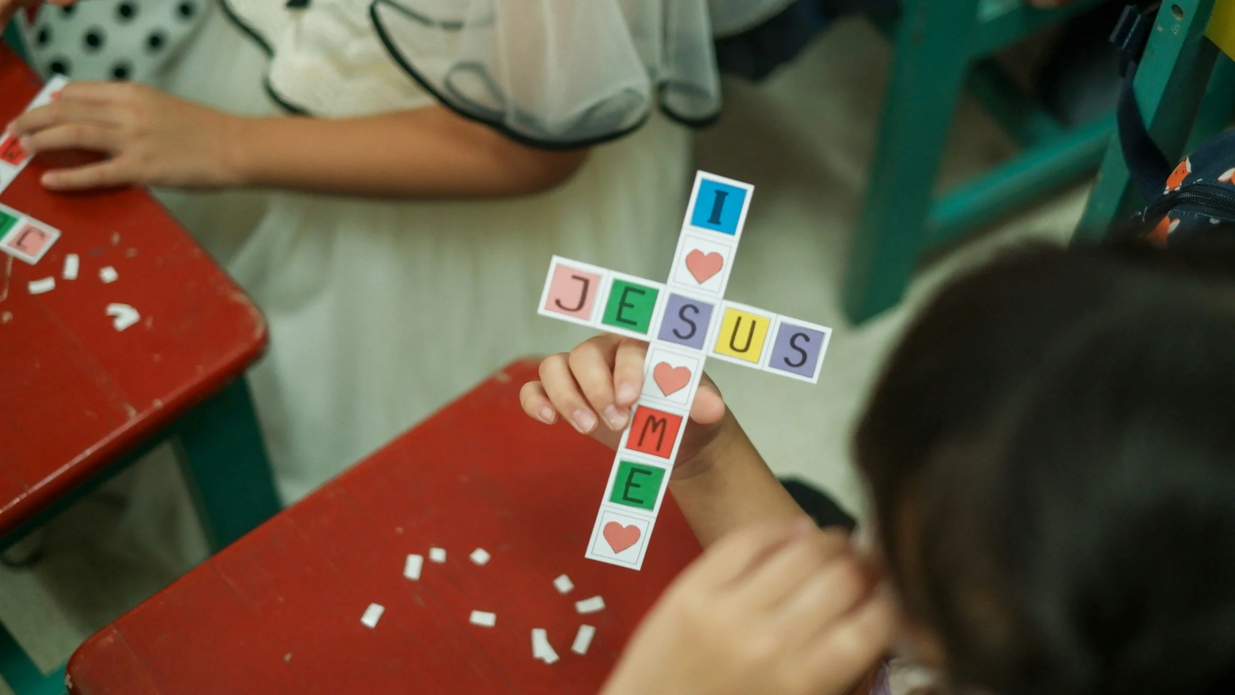A child holding a colorful cross-shaped paper craft spelling 'I LOVE JESUS CHRIST ME' with letter tiles and hearts, sitting at a red desk with scattered small white paper pieces, in a classroom setting.