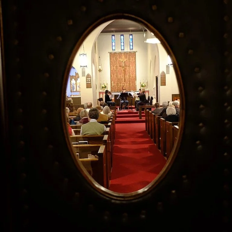 View through a small oval window showing a church interior with people attending a service, musicians playing at the front, and an altar decorated with flowers.