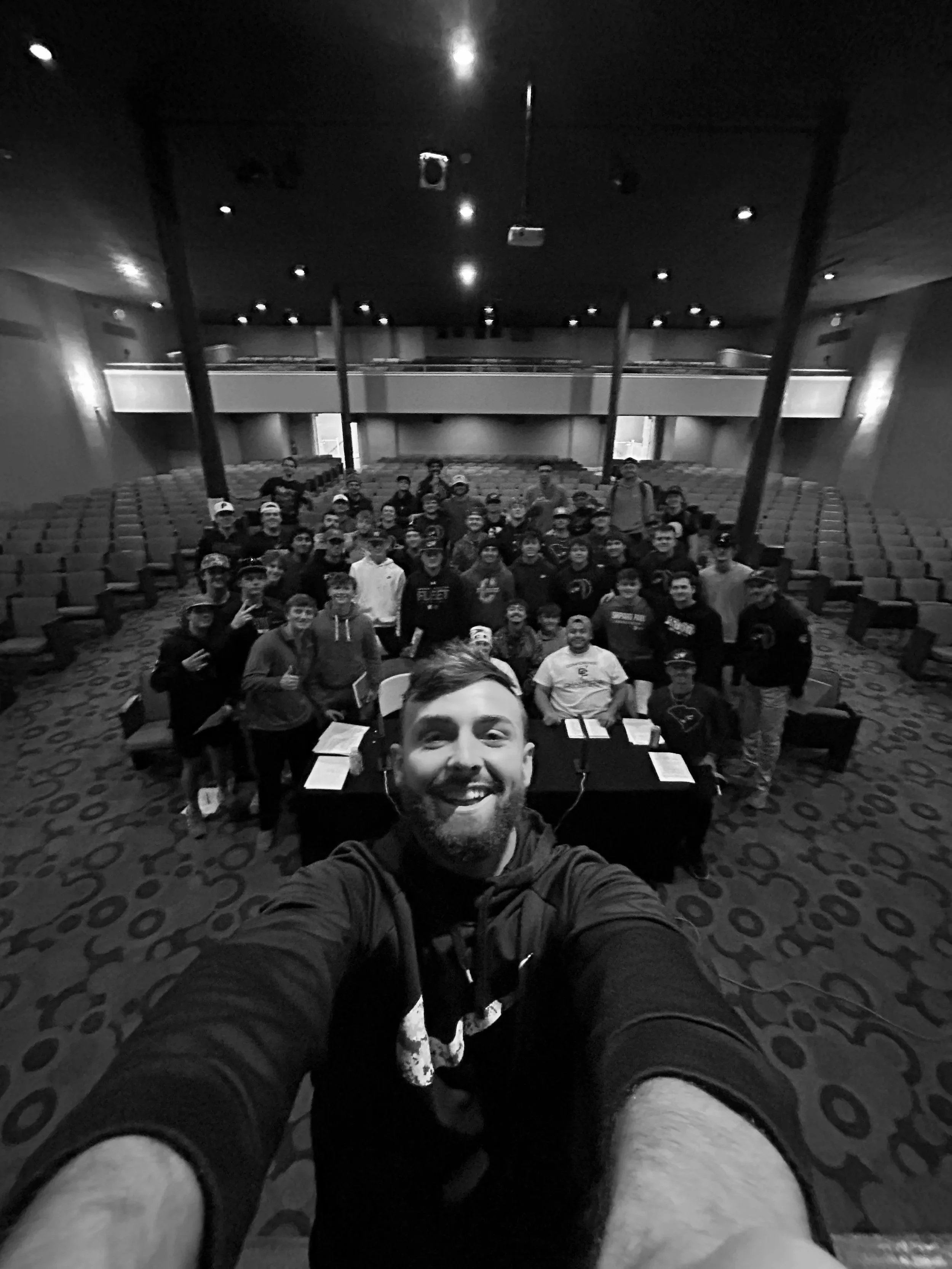 Selfie of a man smiling in front of a group of people seated at a conference table inside an auditorium.