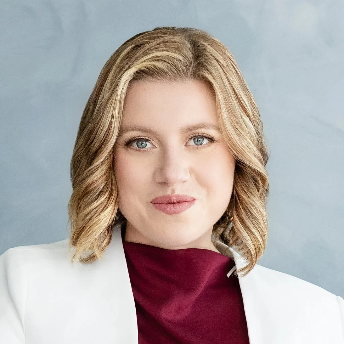 A woman with shoulder-length blonde hair and blue eyes, wearing a white blazer over a maroon top, looking directly at the camera with a neutral expression.