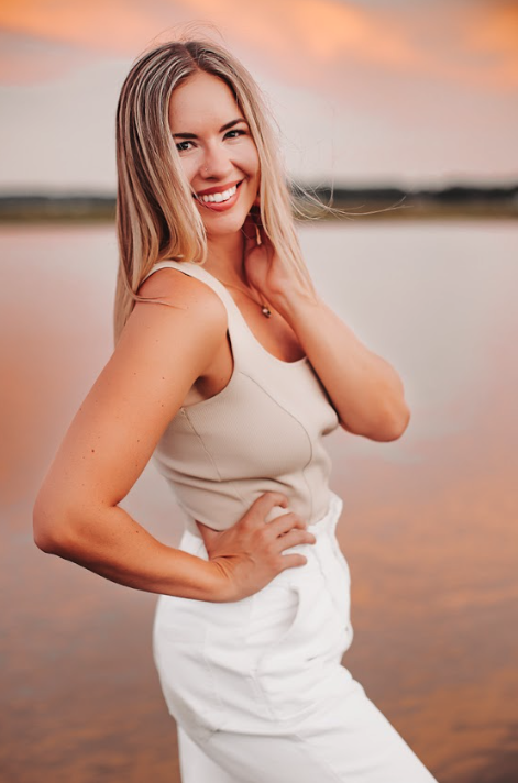 A smiling woman with blonde hair in a sleeveless beige top and white pants, standing outdoors near a body of water during sunset, posing with her hand on her hip.