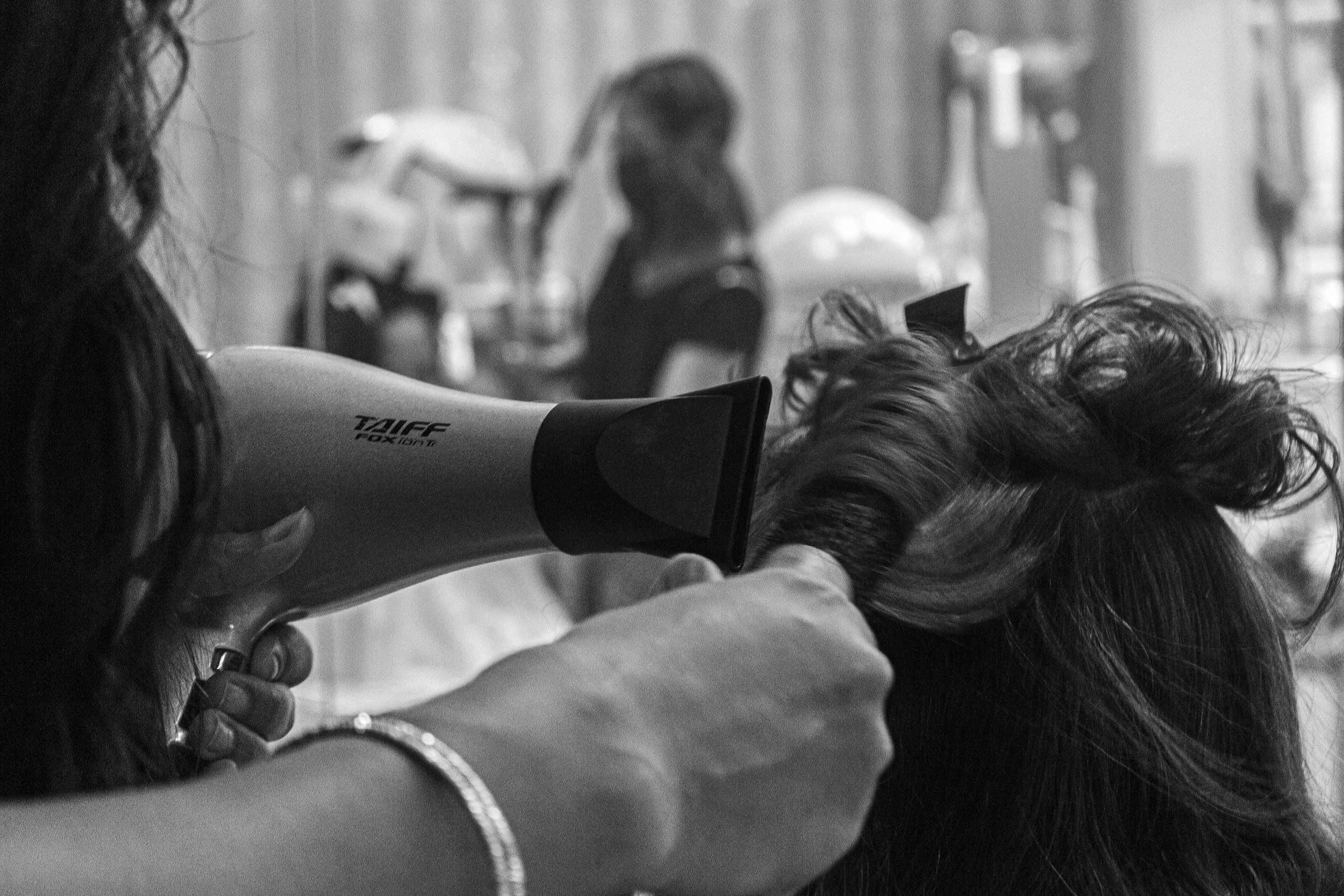 A person using a blow dryer to style a woman's hair in a salon, with another person in the background.