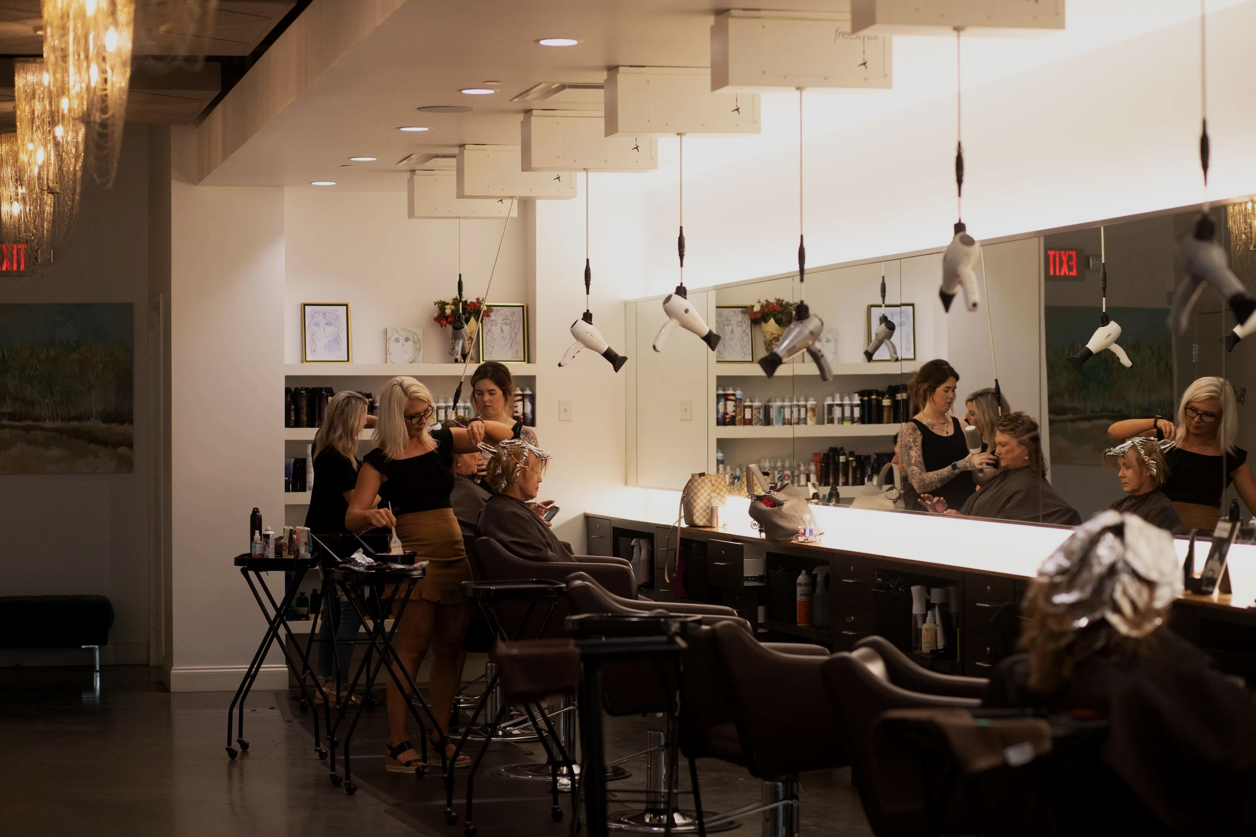 Women getting their hair styled and blow-dried at a salon with hair dryers hanging from the ceiling, shelves with hair products, and framed artwork on the walls.