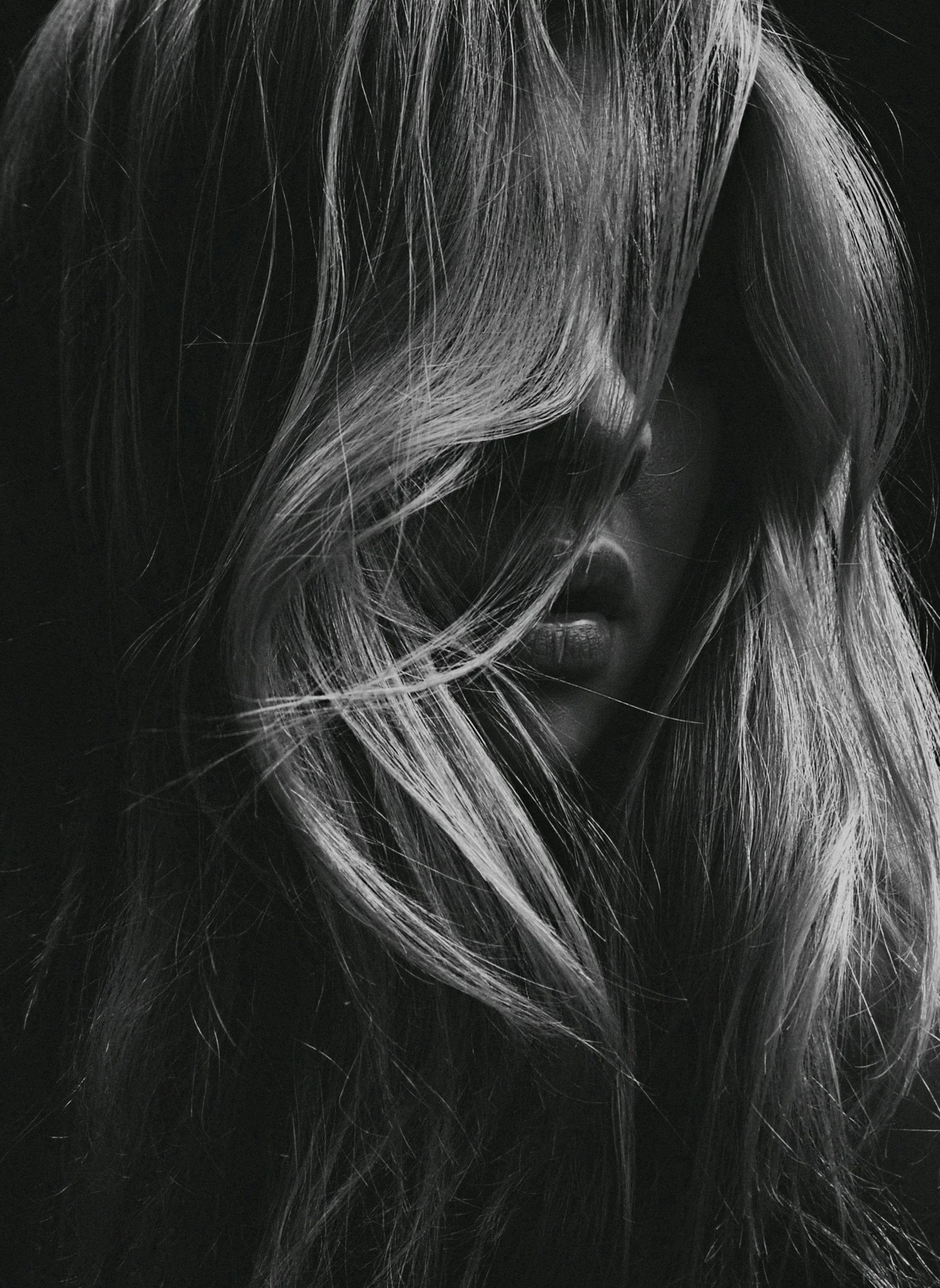 Close-up black and white portrait of a woman with long, wavy hair covering part of her face and lips barely visible.