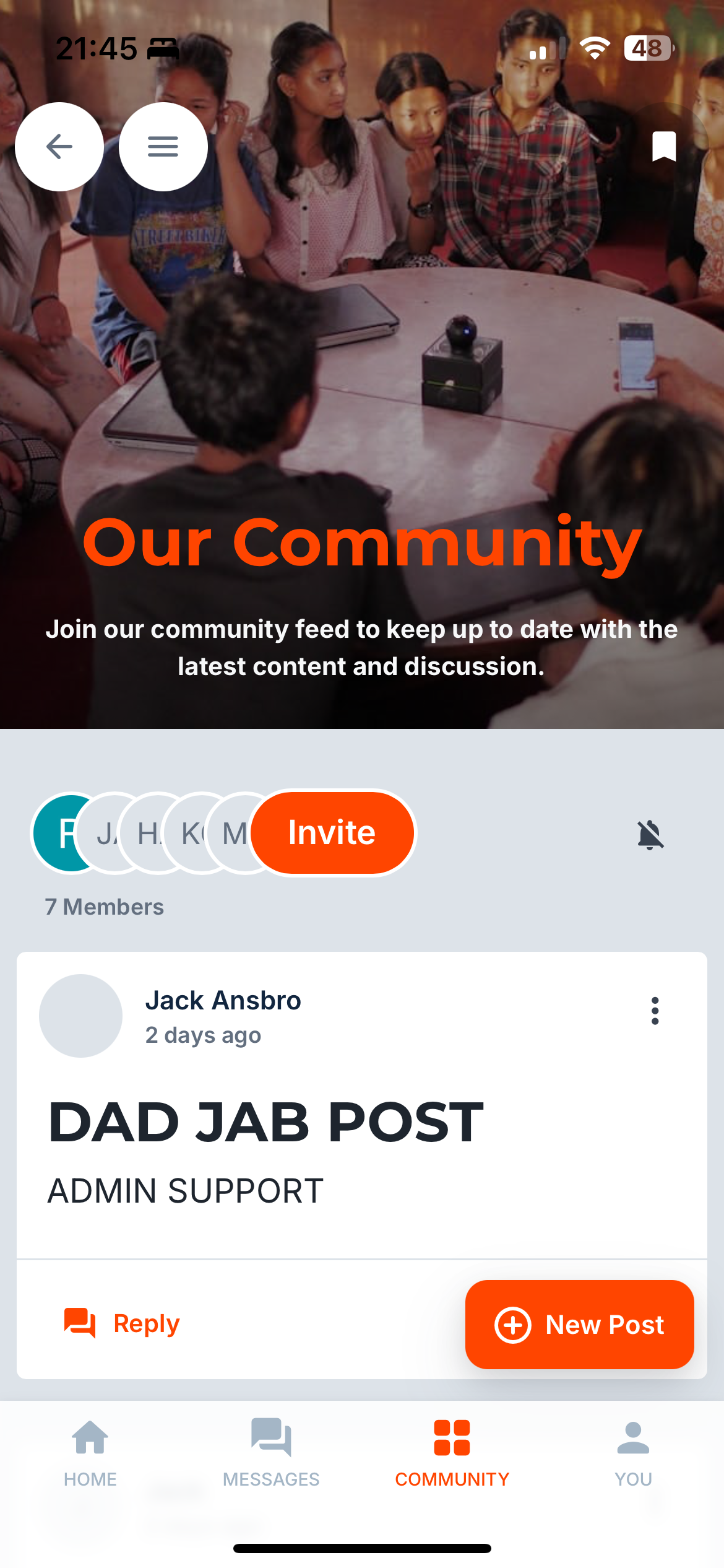 A group of children and a woman are gathered around a table engaging in a discussion, with a small black interactive device on the table. One person is holding a phone, possibly leading the activity.