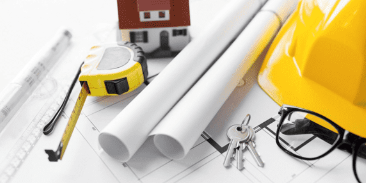 Construction planning tools including a yellow tape measure, rolled blueprints, a yellow hard hat, a small model house, a set of keys, safety glasses, and a ruler on architectural drawings.