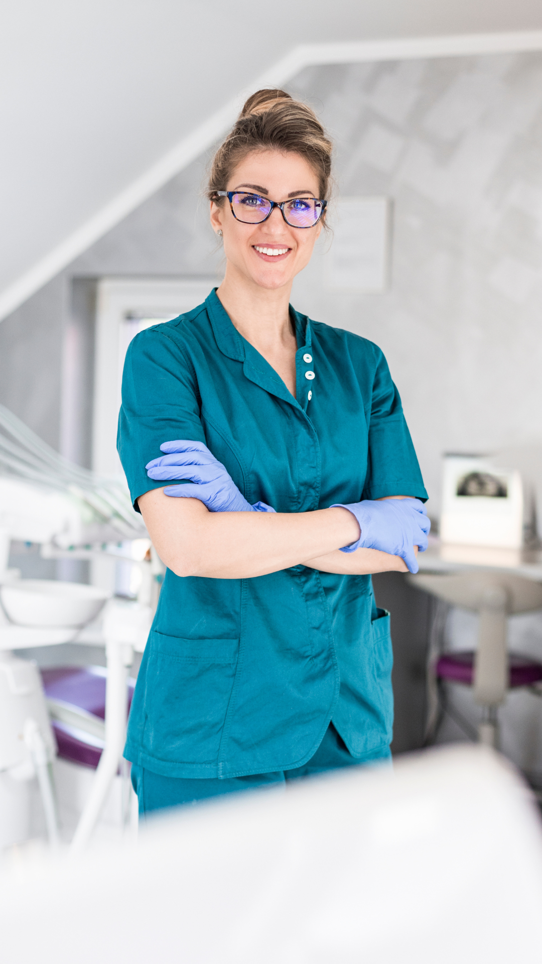 A smiling female healthcare professional wearing glasses, teal scrubs, and purple gloves, standing in a modern medical setting with medical equipment and a window in the background.
