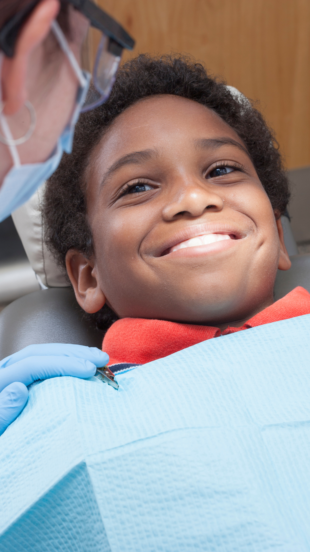 A young boy smiling while sitting in a dental chair, wearing a red shirt and a dental bib, with a dentist inspecting his teeth.