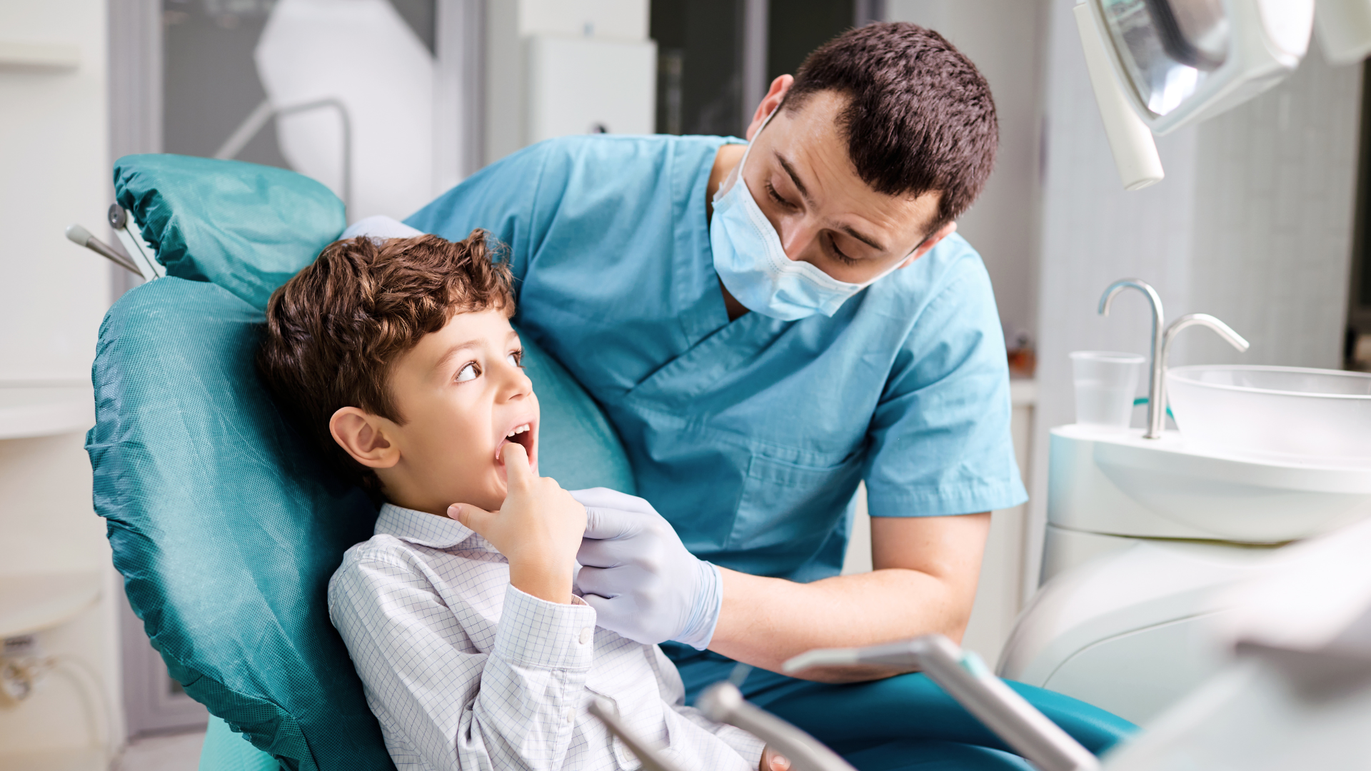 A young boy sitting in a dental chair with a caregiver. The boy appears to be talking or explaining something, with a curious or concerned expression. The caregiver, wearing dental scrubs, a face mask, and gloves, leans in attentively while listening to him. The dental office setting includes dental equipment and a sink in the background.