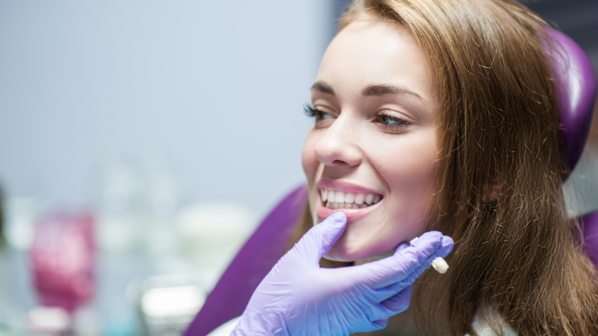 A young woman at a dental clinic is smiling as a dental professional, with gloved hands, examines her teeth.