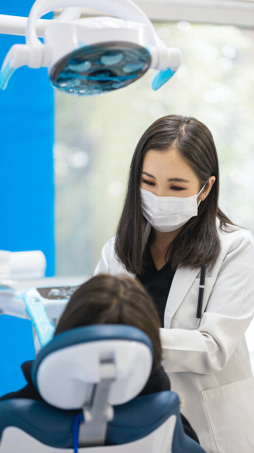 A female dentist or dental hygienist wearing a face mask and white coat cleaning or examining a patient's teeth in a dental office.