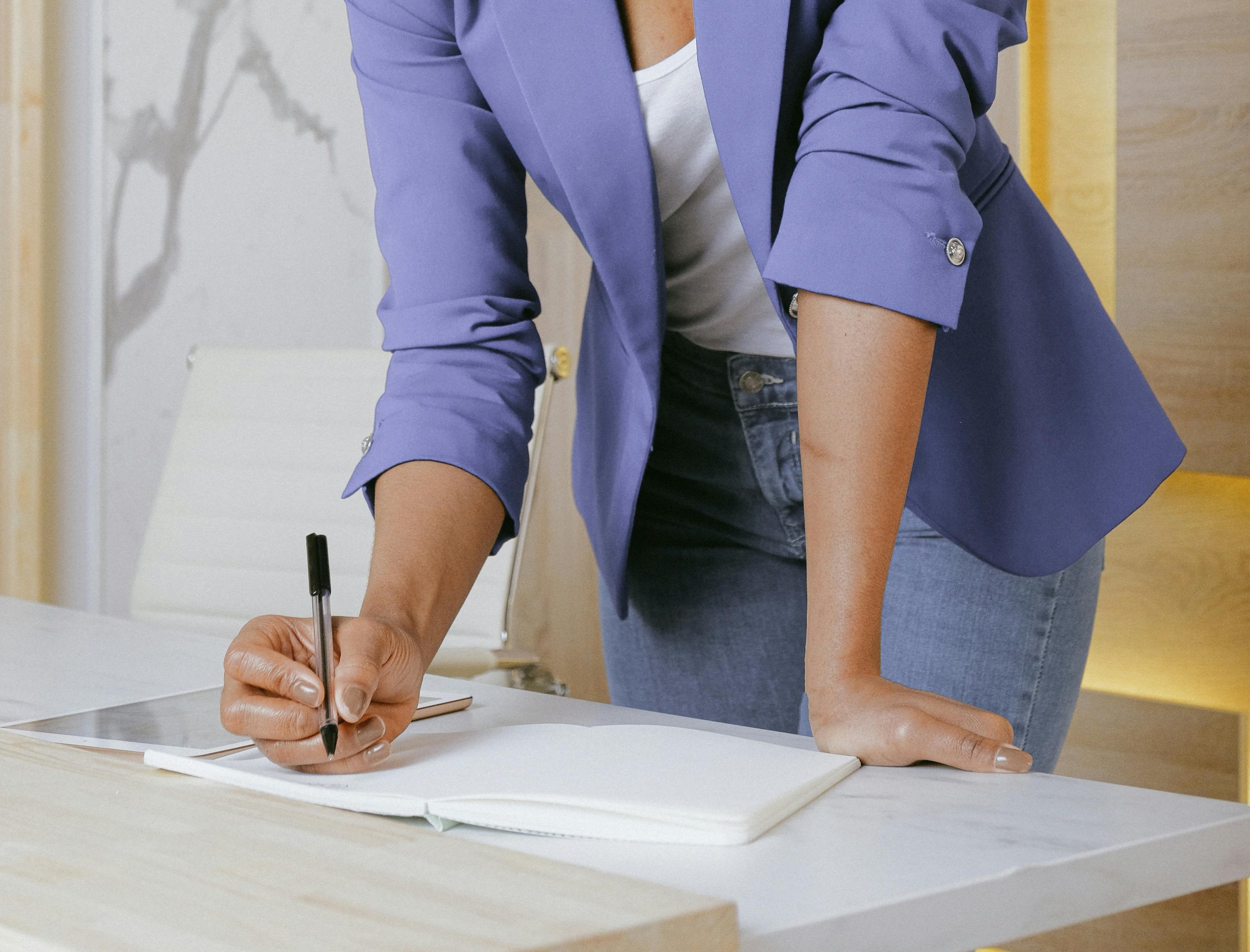 Person in a blue blazer and jeans writing in a notebook with a pen on a desk.