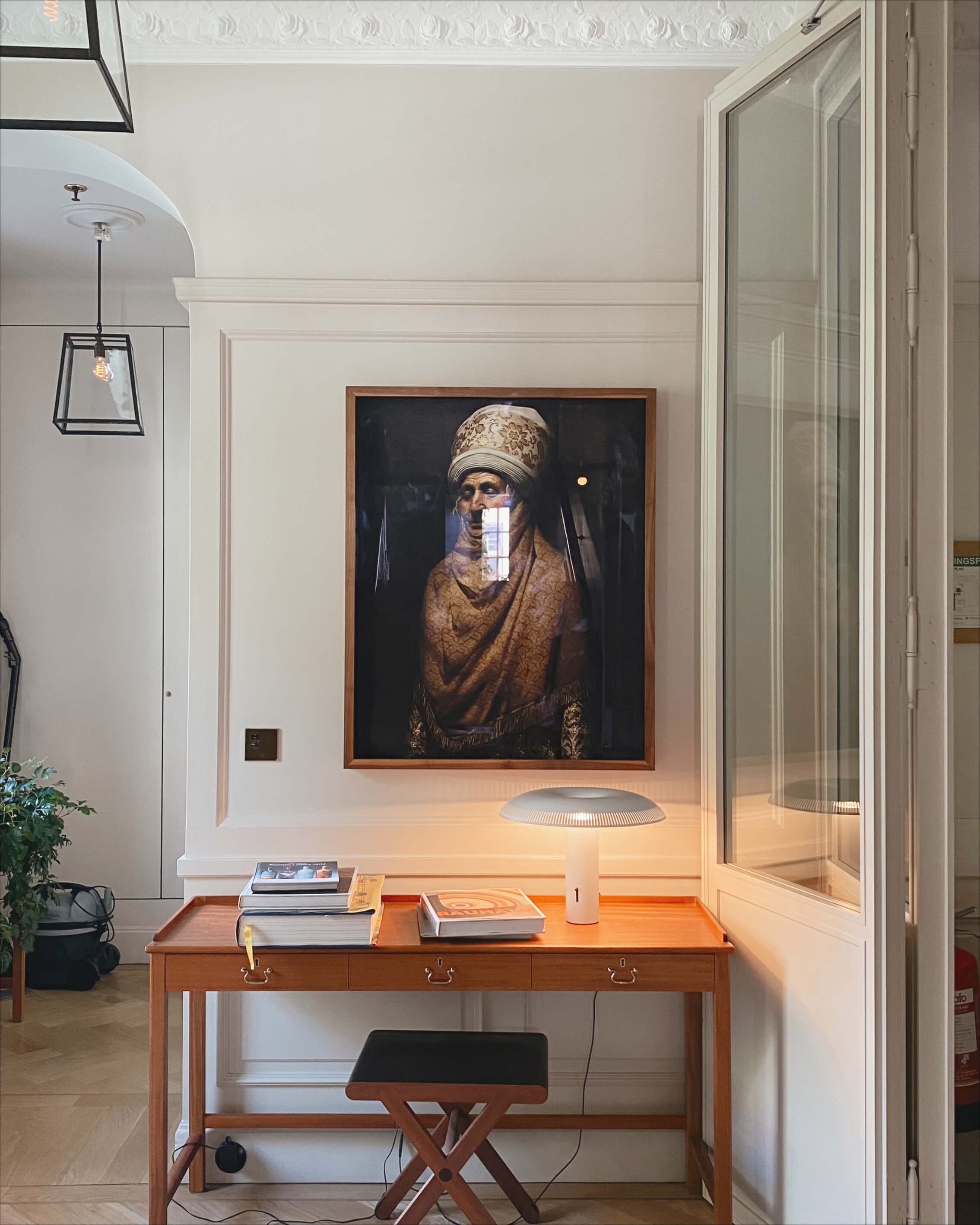 Interior of a room with a desk, a small lamp, books, and a framed portrait of an elderly woman on the wall.