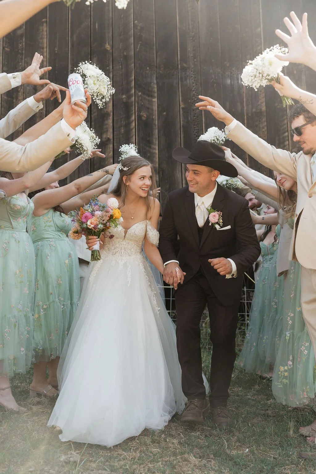 A bride and groom holding hands and walking together at their wedding, surrounded by bridesmaids and groomsmen throwing flower petals and holding bouquets, in front of a wooden fence.