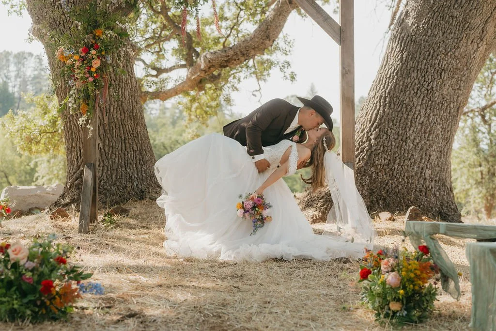 Wedding scene with a bride and groom kissing under a tree, surrounded by flowers.