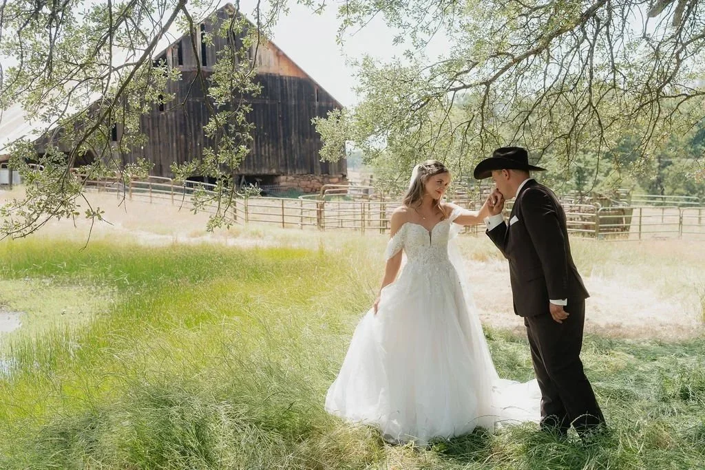 A bride and groom share a tender moment outdoors, with the groom kissing the bride's hand. They are dressed in wedding attire, with the bride in a white gown and the groom in a black suit and cowboy hat. A rustic barn and fencing are visible in the background, under a leafy tree.