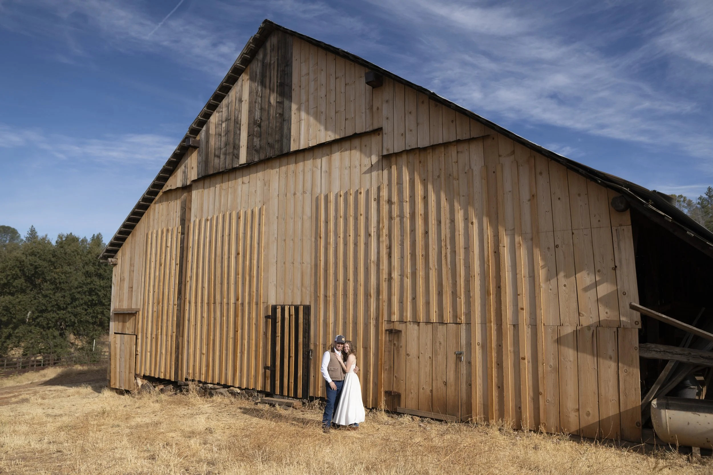 A man and woman standing together outside a large wooden barn with a partly cloudy blue sky in the background.
