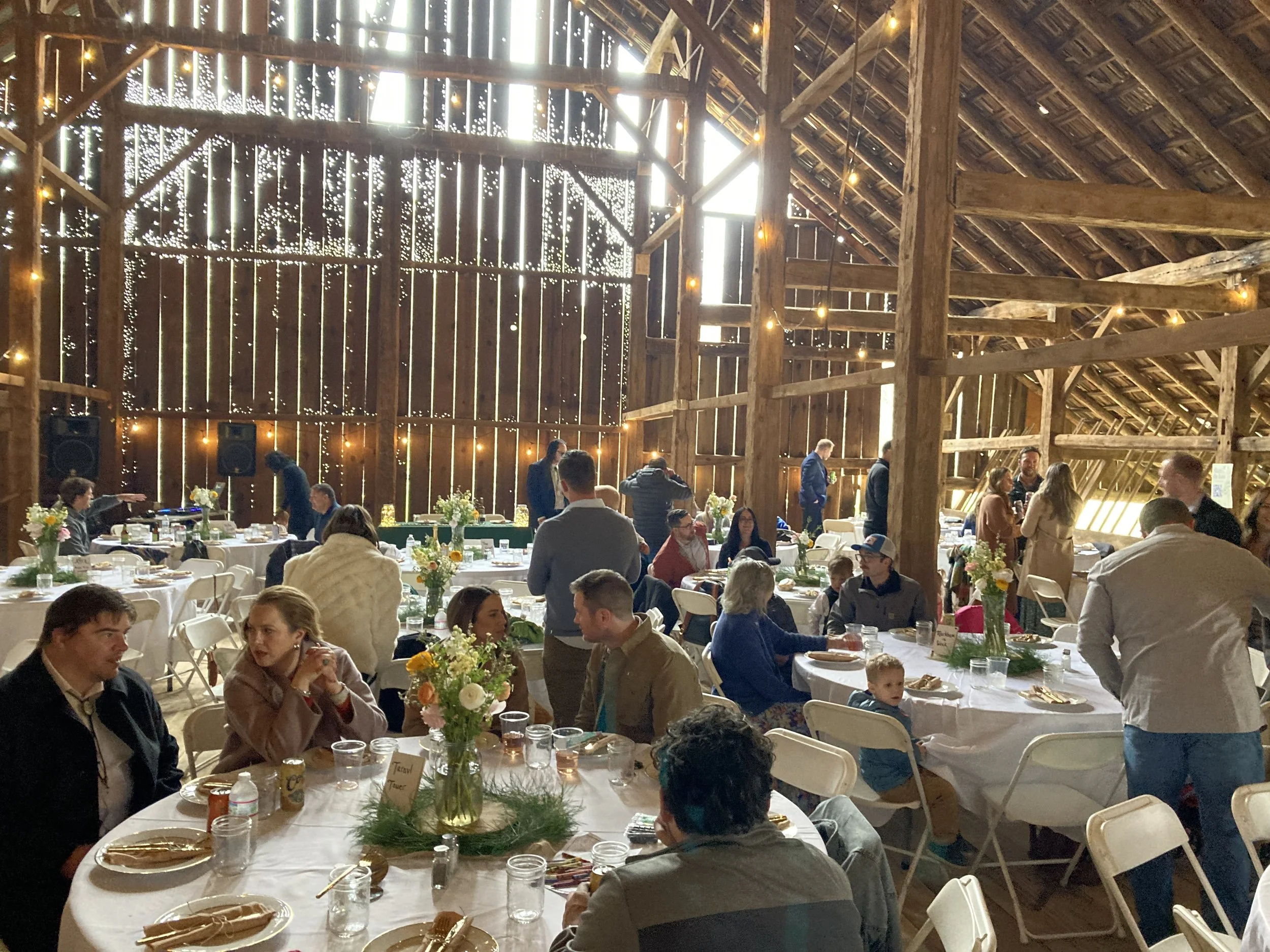People gathered in a large rustic barn decorated for a social event, with round tables covered in white tablecloths, floral centerpieces, and string lights hanging from the wooden beams.