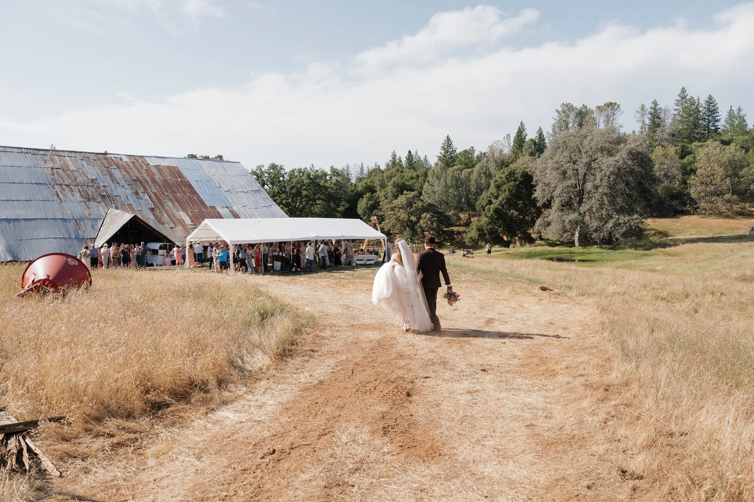 Bride and groom walking on a dirt path towards a rustic outdoor wedding ceremony with guests and a large barn in the background.