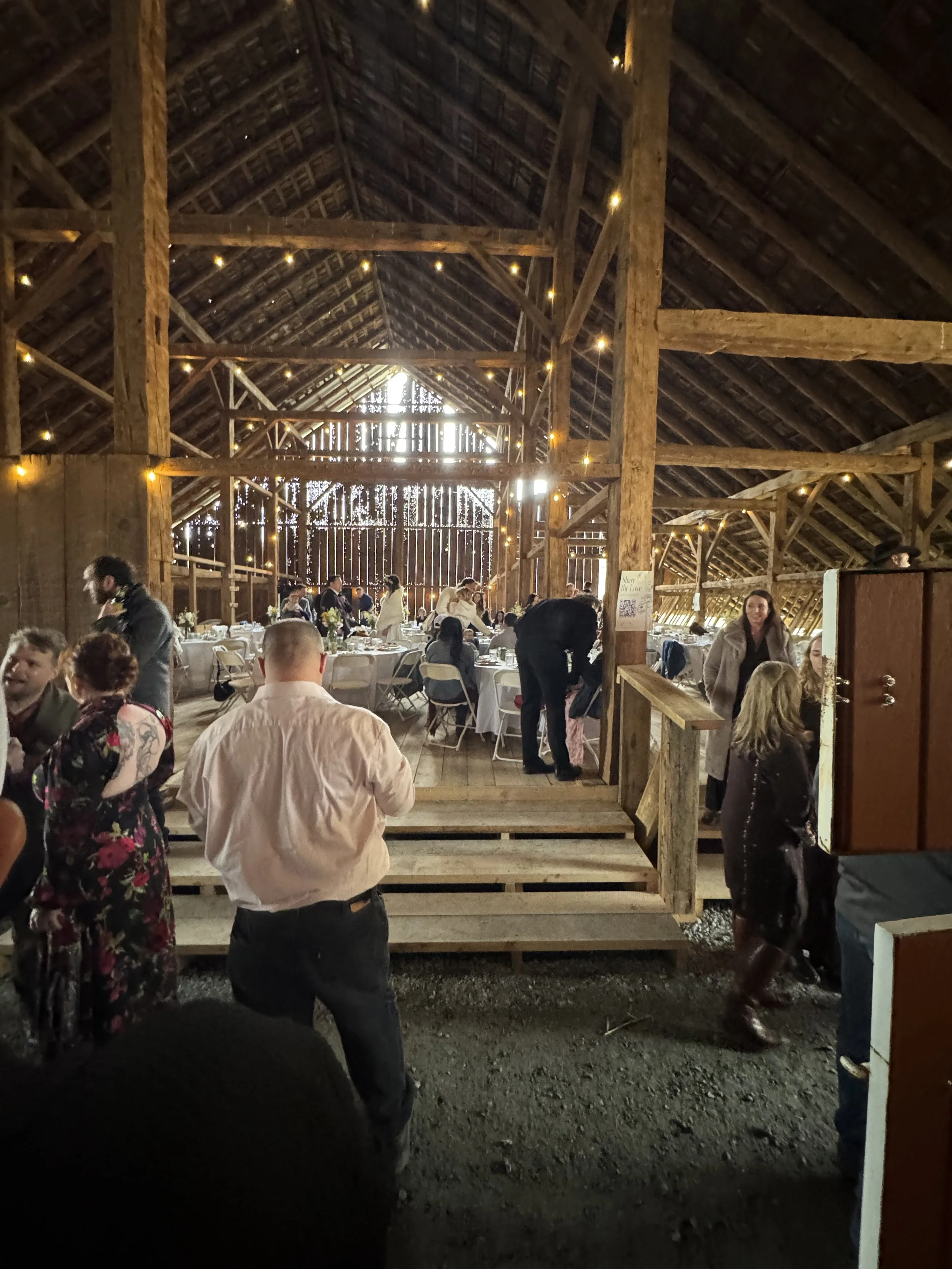People gathering inside a rustic barn with wooden beams, attending a wedding reception with tables and chairs, and string lights hanging across the ceiling.