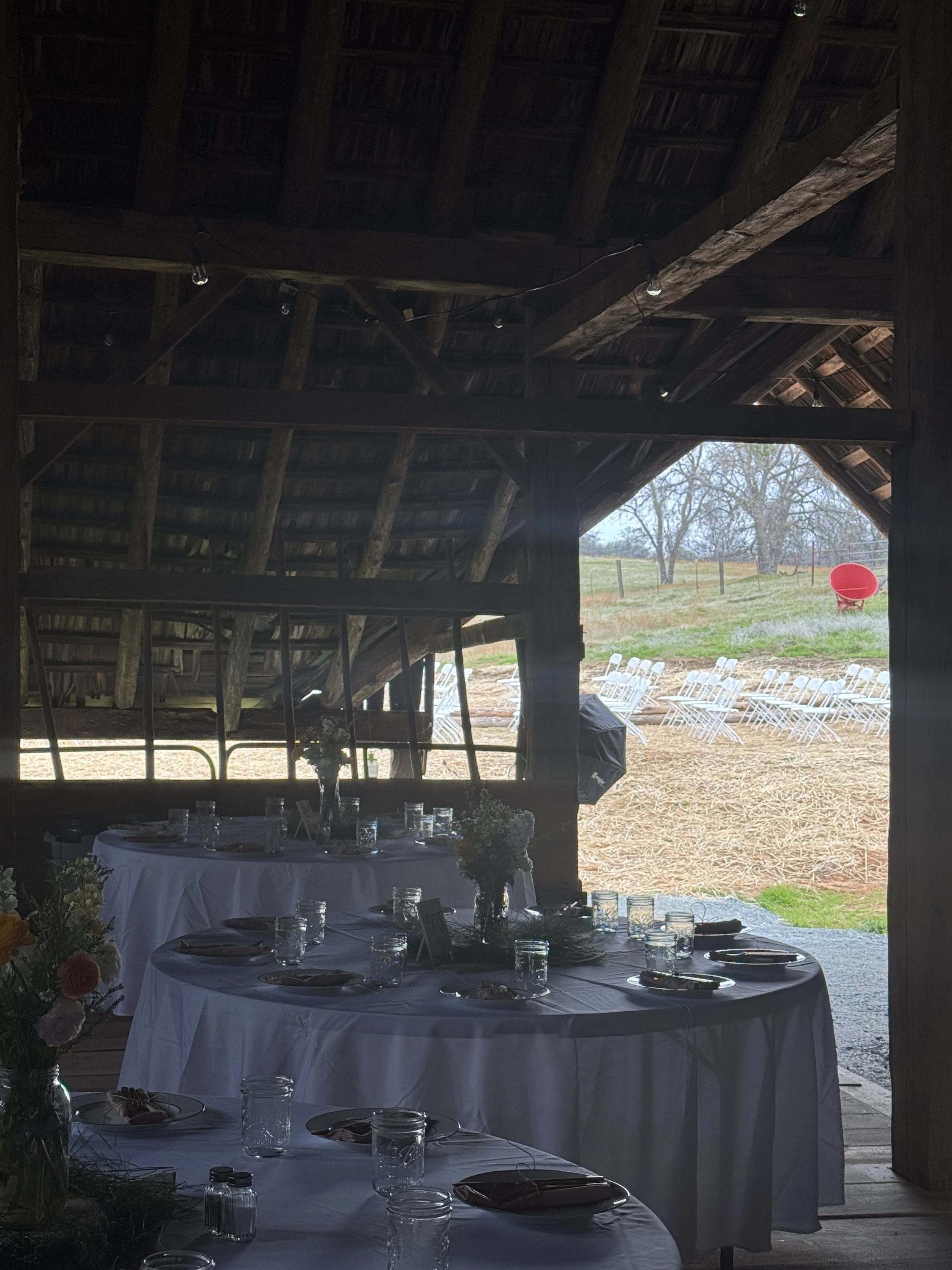 Inside a rustic barn set for an outdoor event with round tables covered in white tablecloths, glassware, and floral centerpieces, with a view of a grassy field and white chairs outside.