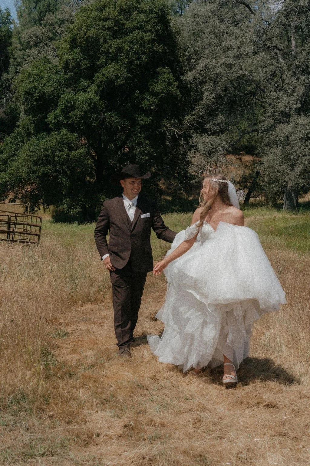 A newlywed couple holding hands and smiling outdoors in a grassy field with trees in the background, the bride wearing a white wedding gown and veiled, and the groom in a dark suit and cowboy hat.