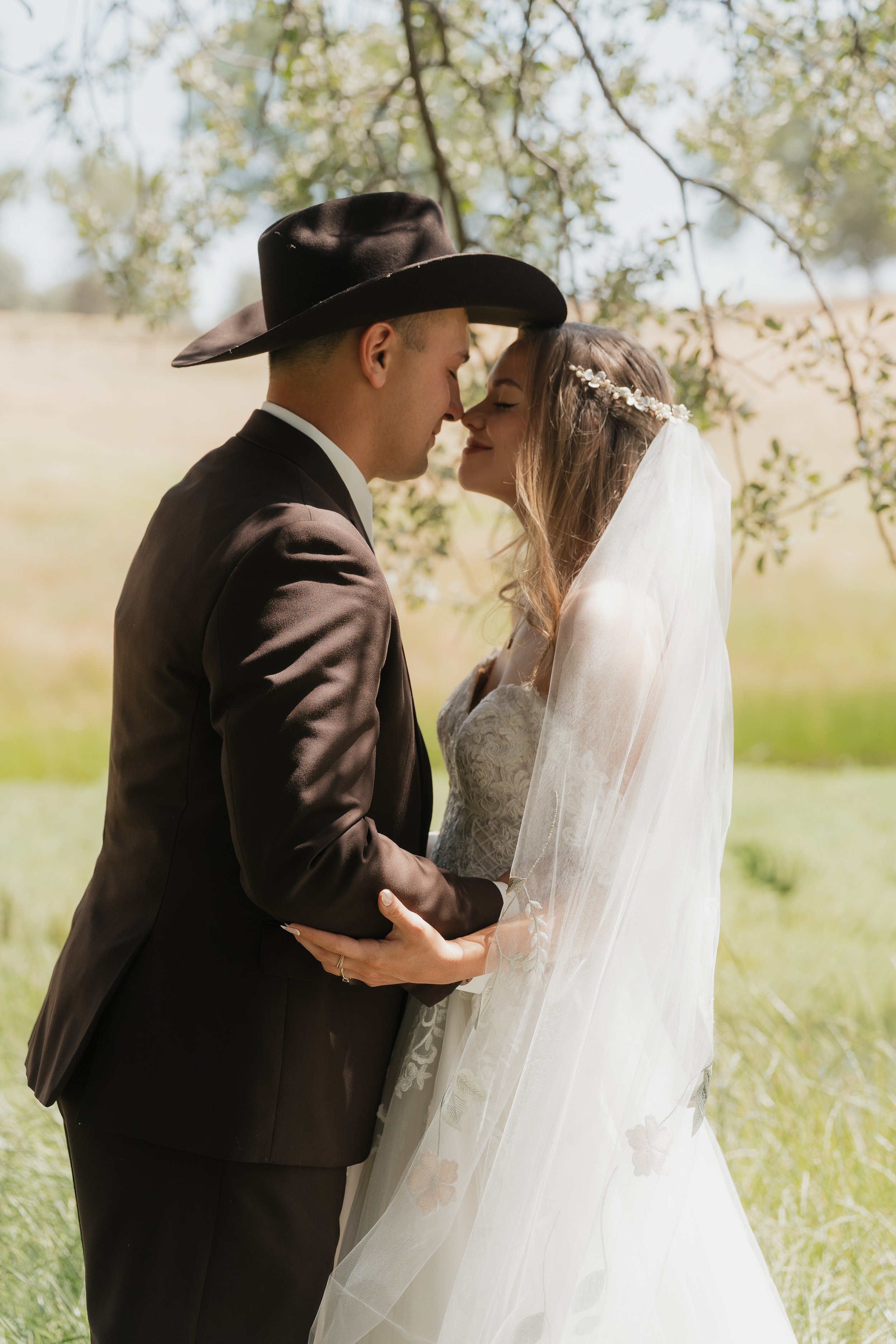 A bride and groom stand close together outdoors, touching foreheads and holding hands, with trees and soft sunlight in the background.