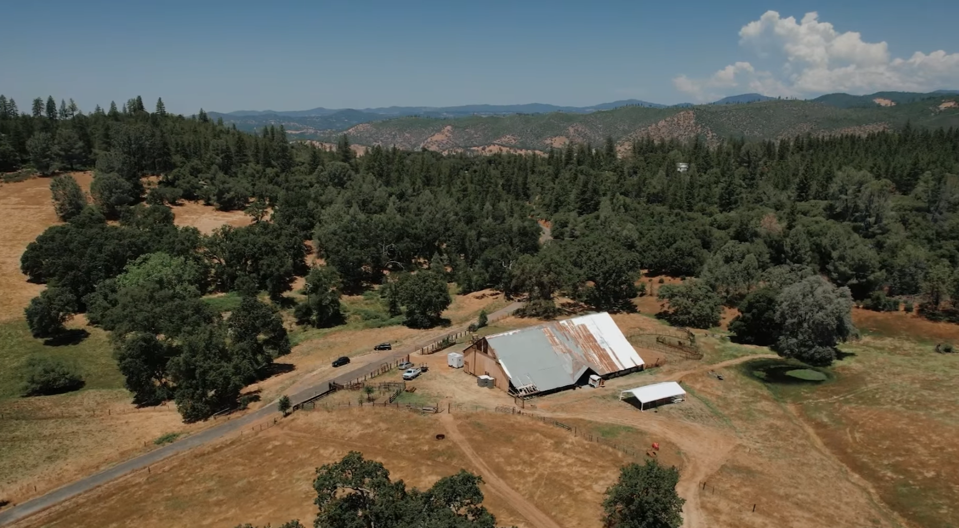 Overhead view of a rural landscape with a large barn, a small shed, a pond, and a few parked cars, surrounded by trees and hills under a partly cloudy sky.
