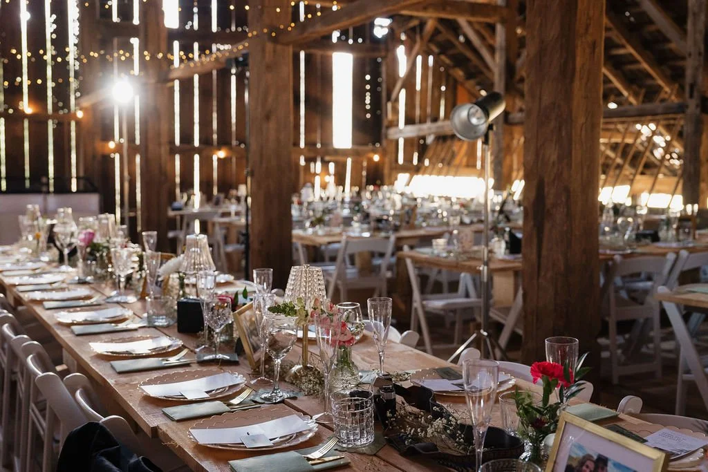 Long wooden banquet table set with white plates, silverware, glassware, floral arrangements, and decorated with candles inside a rustic barn with wooden beams and string lights.
