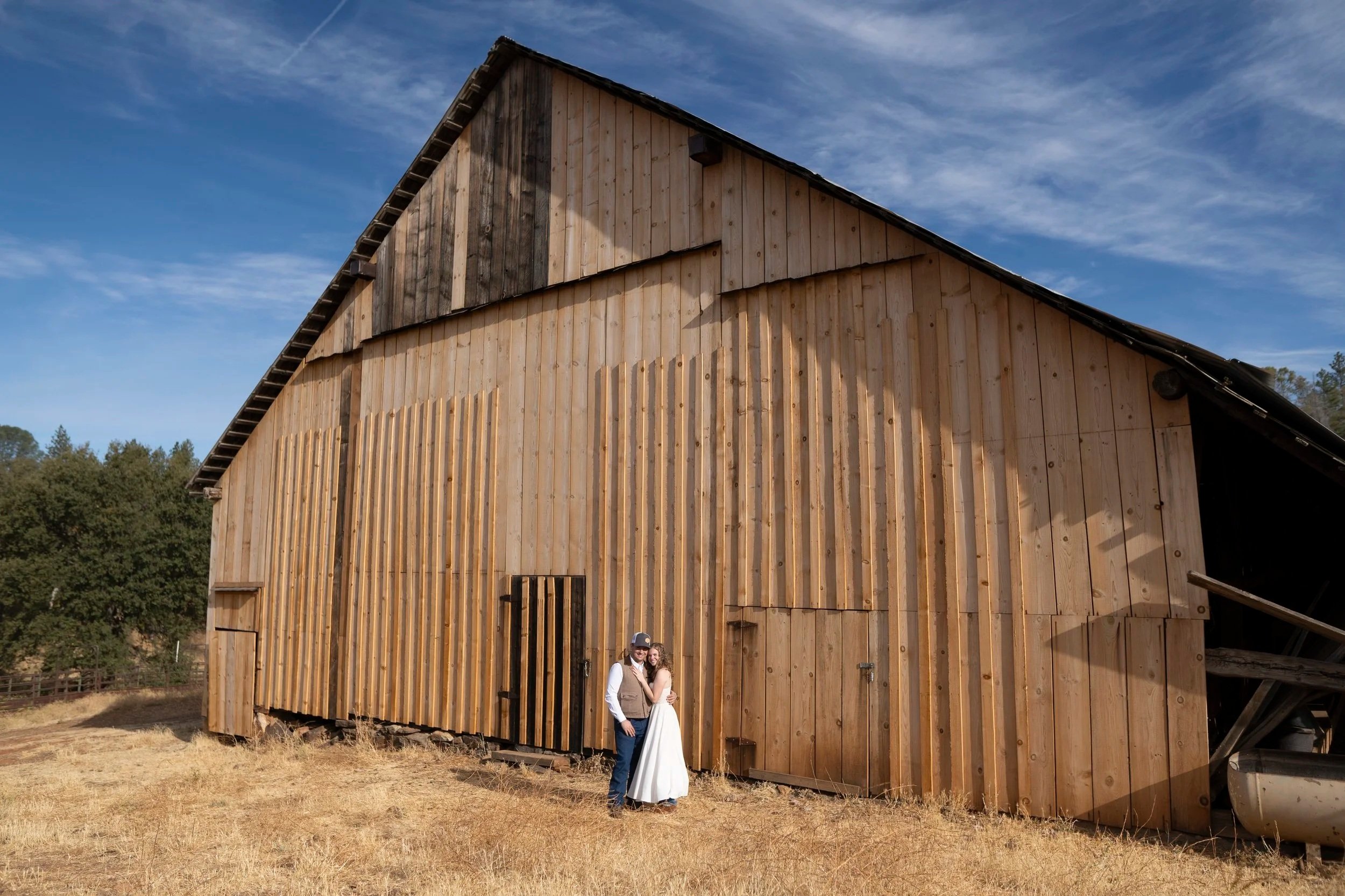 A couple stands arm in arm outside a large wooden barn with a bright blue sky and clouds overhead.