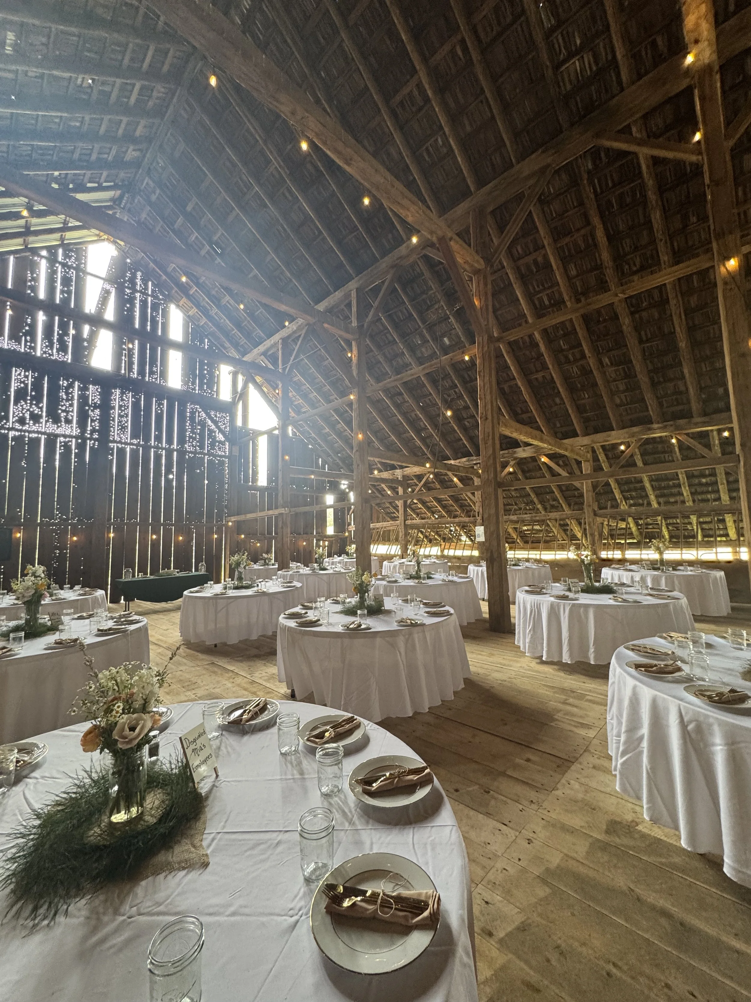 Wedding reception setup in a rustic barn with round tables covered in white tablecloths, floral centerpieces, and place settings, with wooden beams and walls illuminated by string lights.