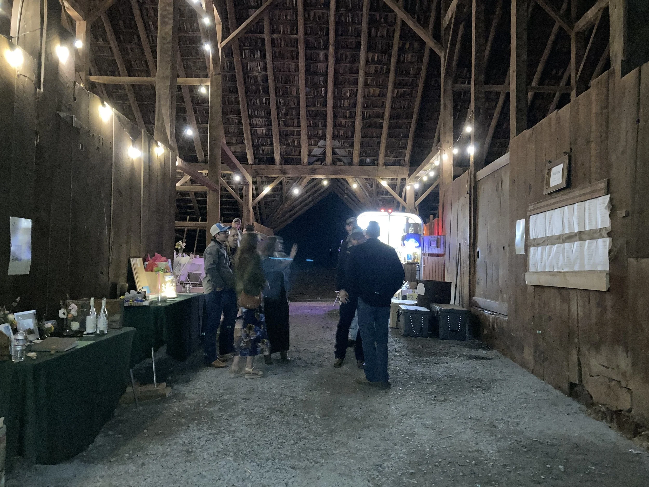 People gathered inside a rustic barn with string lights, near a food or drink stand, at nighttime.