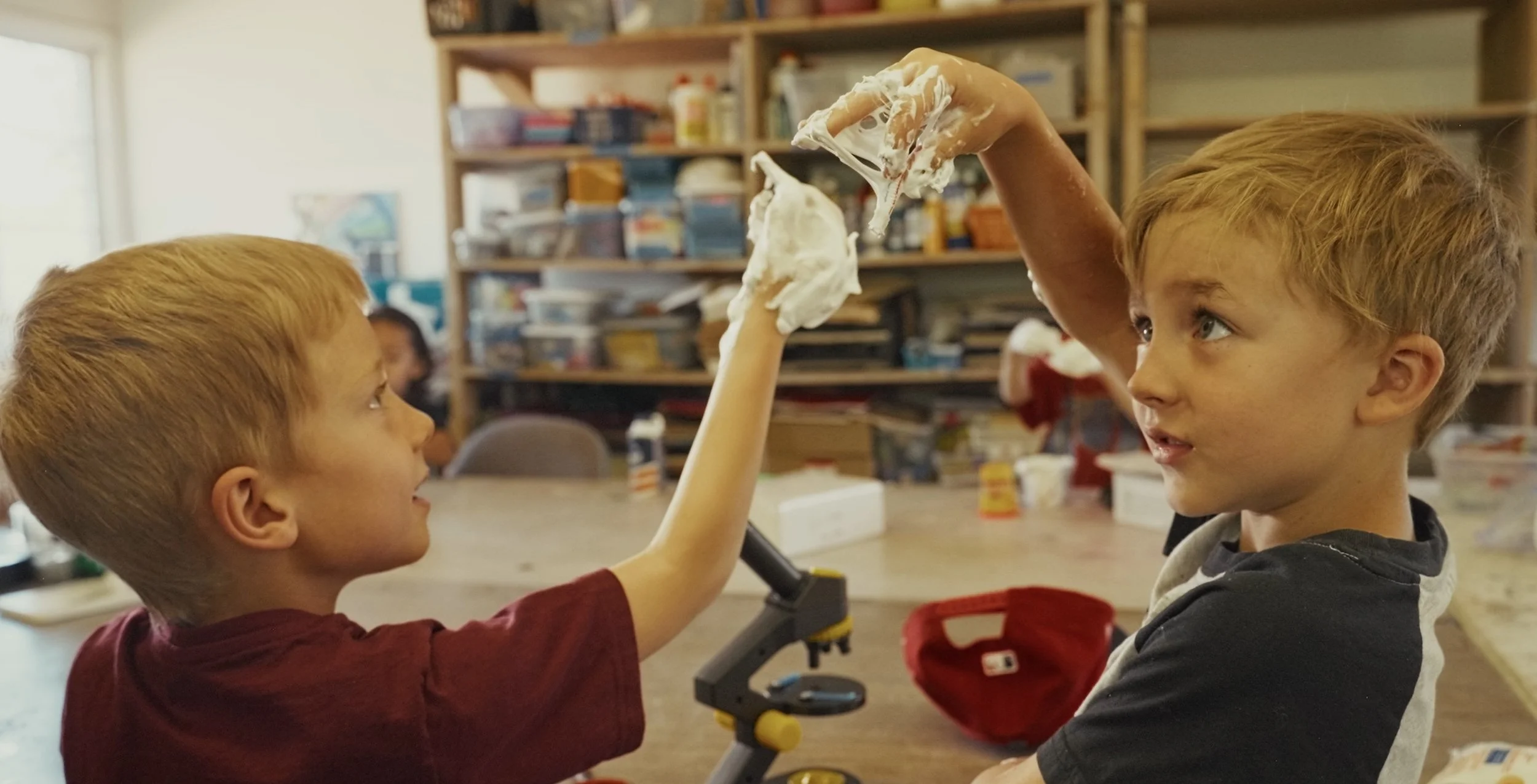 Two young boys in a Meteor Crater Education Alliance summer camp in Flagstaff, with one boy on the left holding up a cloud of foam or marshmallow, and the other boy on the right watching with interest, with shelves and supplies in the background.