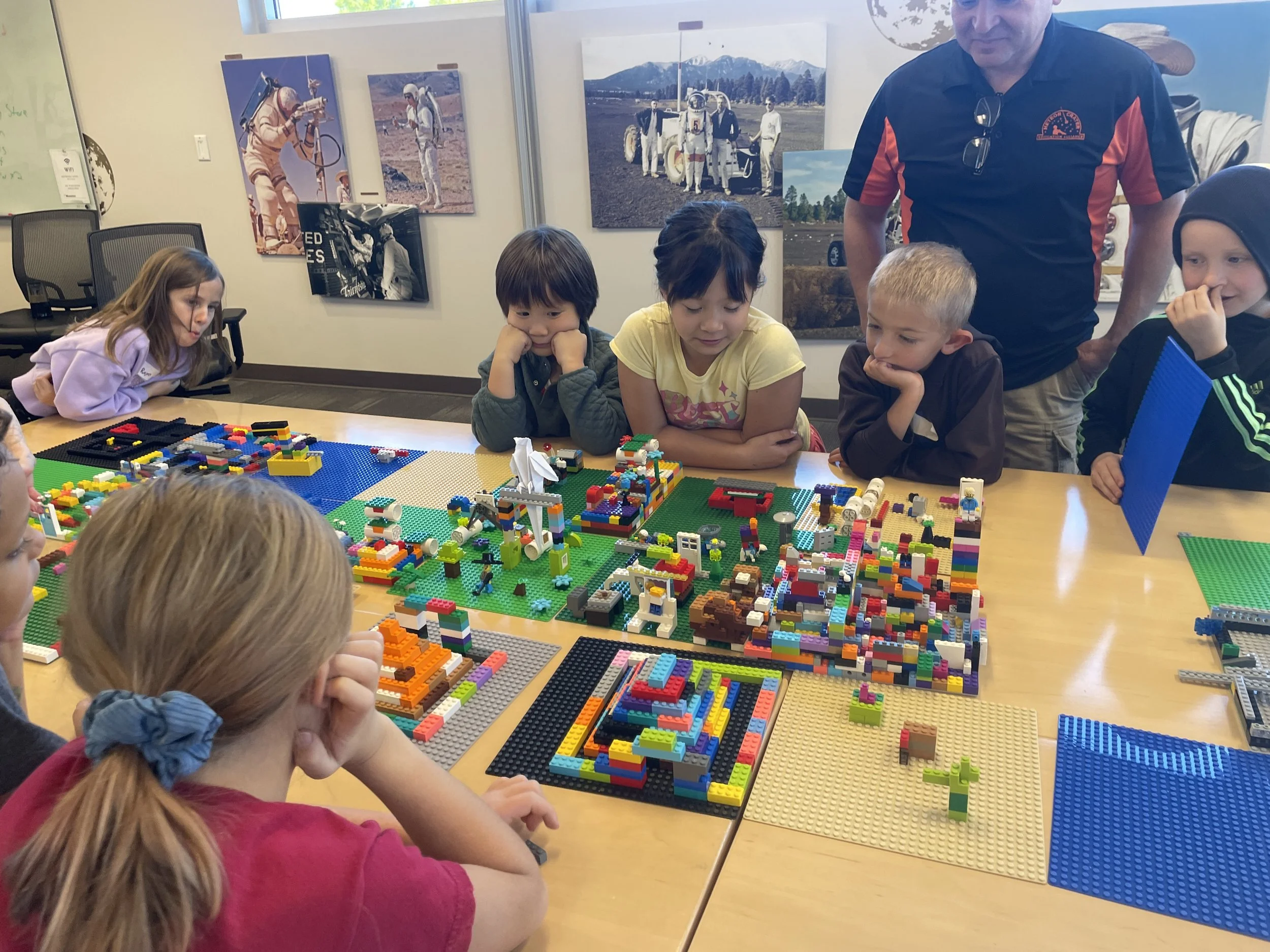 Children and an adult gathered around a table with colorful LEGO structures in a room decorated with space and astronaut posters. They are exploring science and STEM projects in a summer camp in Flagstaff.