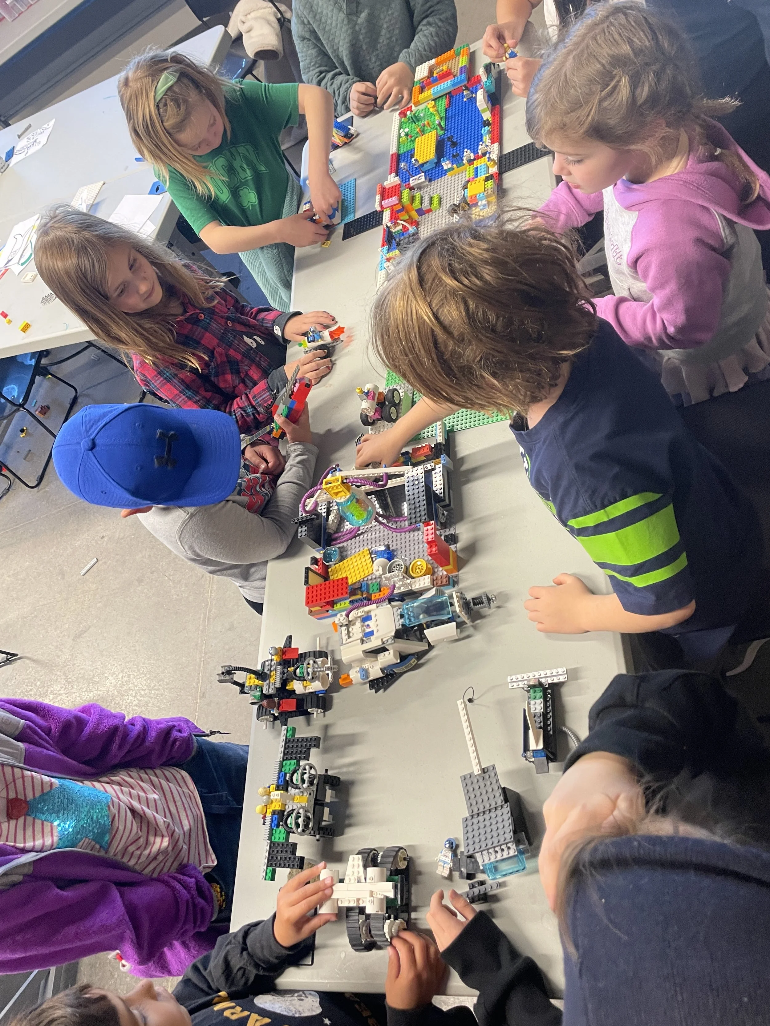 Children building and playing with LEGO bricks on a table in a classroom or recreational setting.