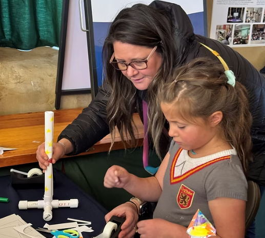 A woman and a young girl working together on a craft project at a table, with craft supplies like tape, scissors, and paper around them.