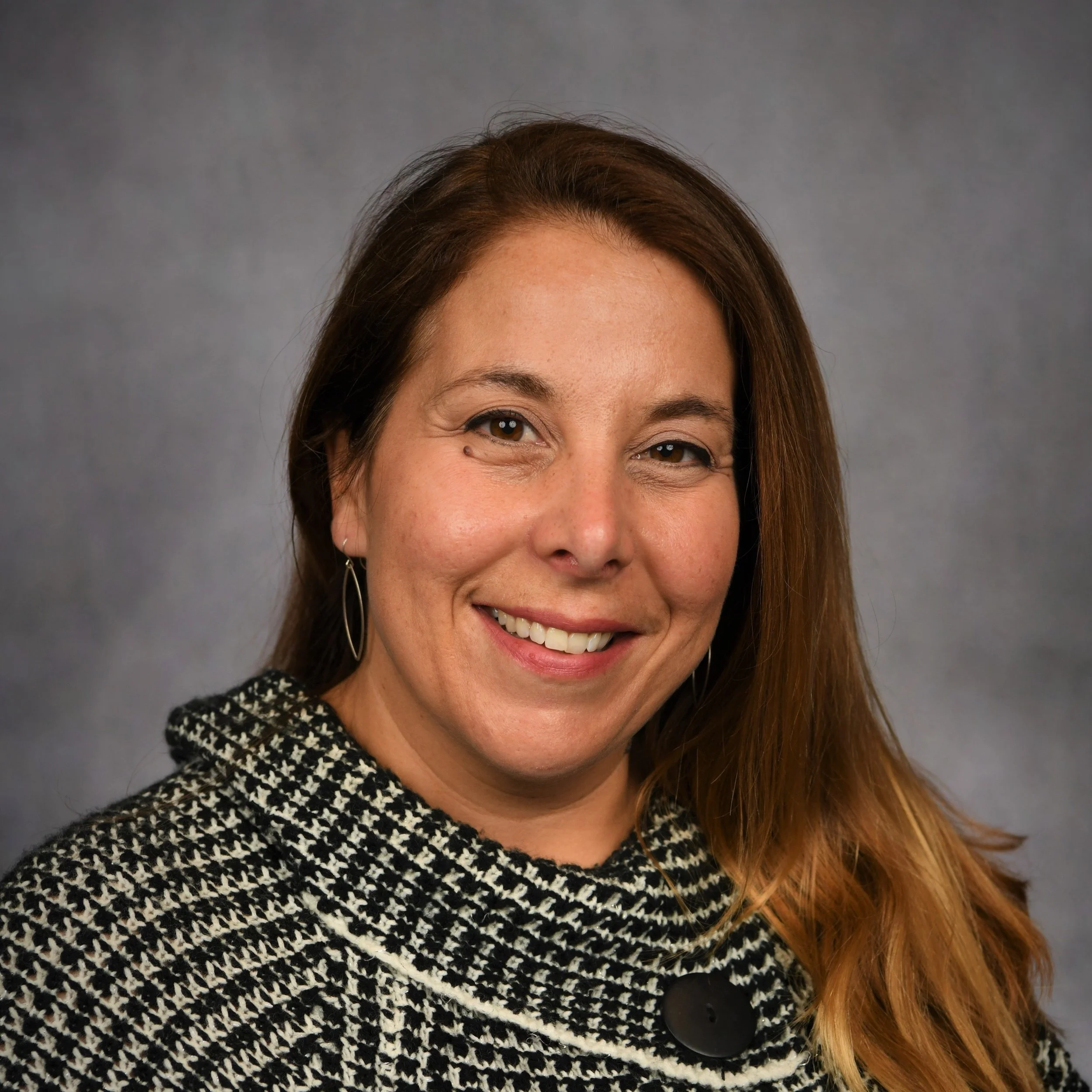 Headshot of a smiling woman with long brown hair, wearing a patterned black and white jacket with a large black button, and silver earrings, against a gray background.