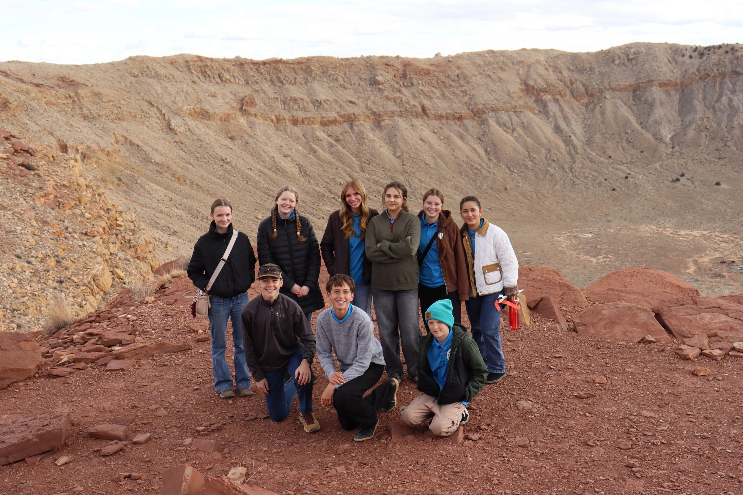Group of ten young people outdoors on rocky terrain with red and beige hills in the background, some kneeling and some standing, dressed in casual outdoor clothing.