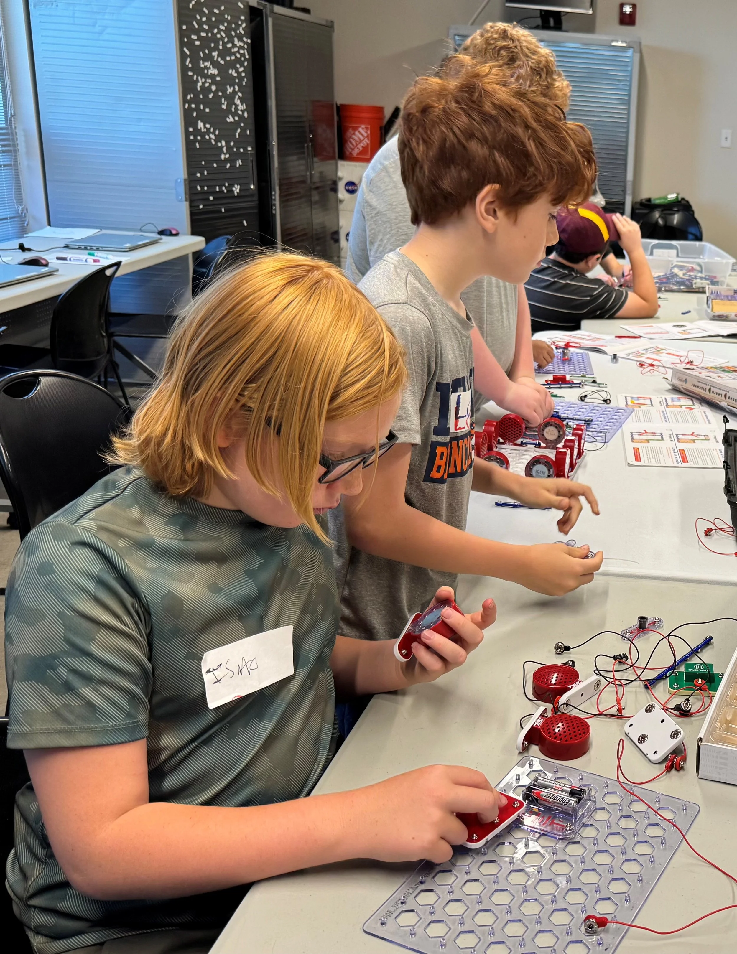Children participating in a STEM activity, working with electronic components and tools on a table in a classroom or workshop setting.