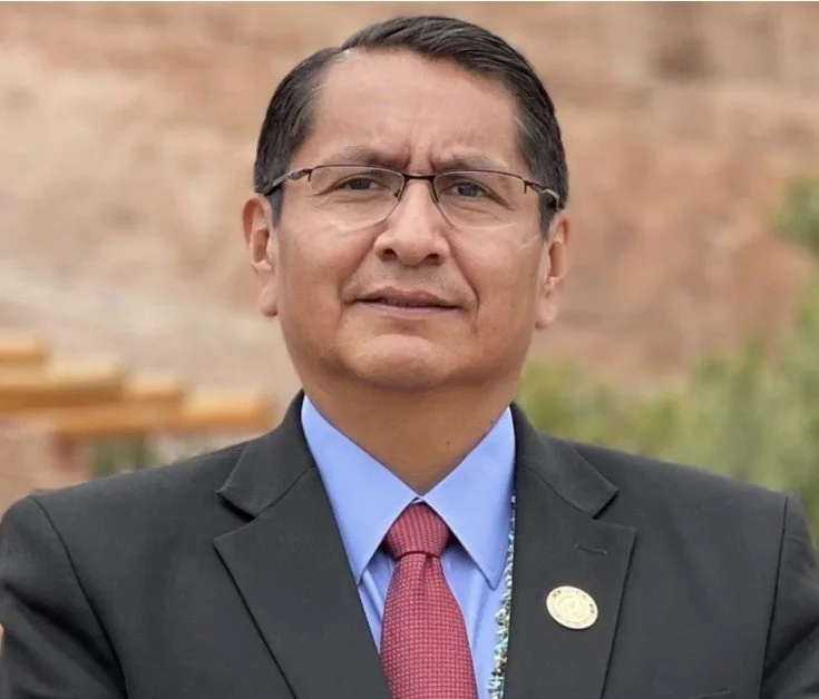 A man in a suit and tie wearing glasses, standing outdoors with a blurred brick wall and some greenery in the background.