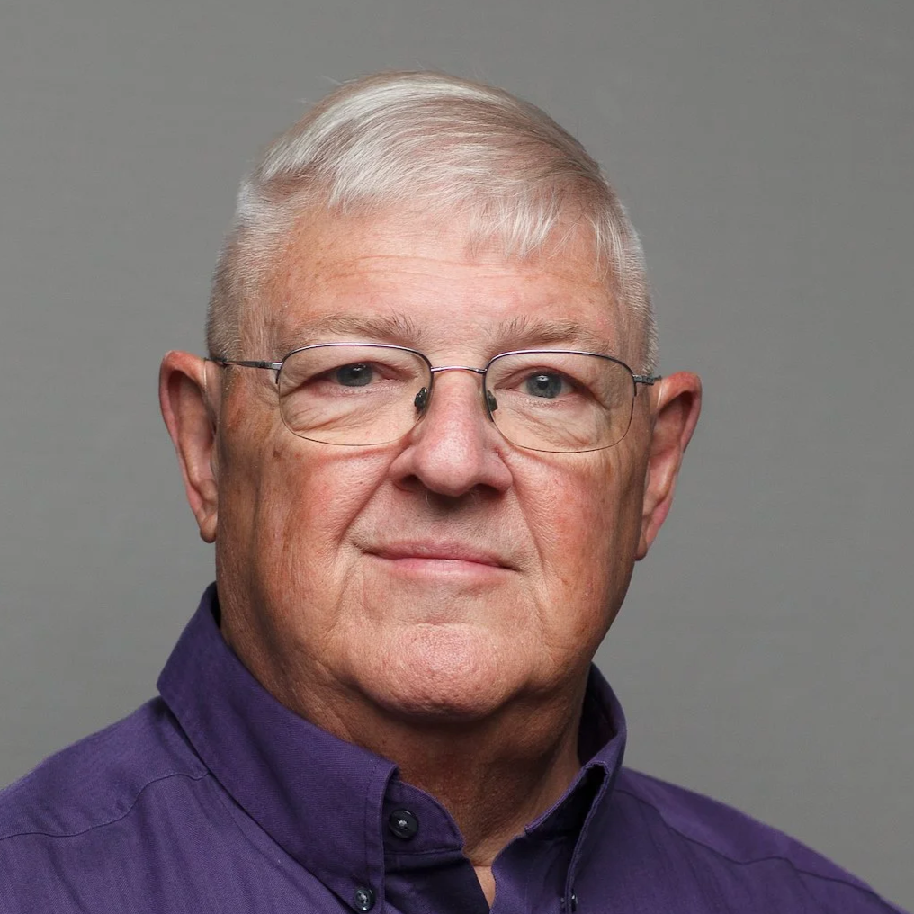 Portrait of an older man with silver hair, glasses, and wearing a purple shirt against a plain gray background.