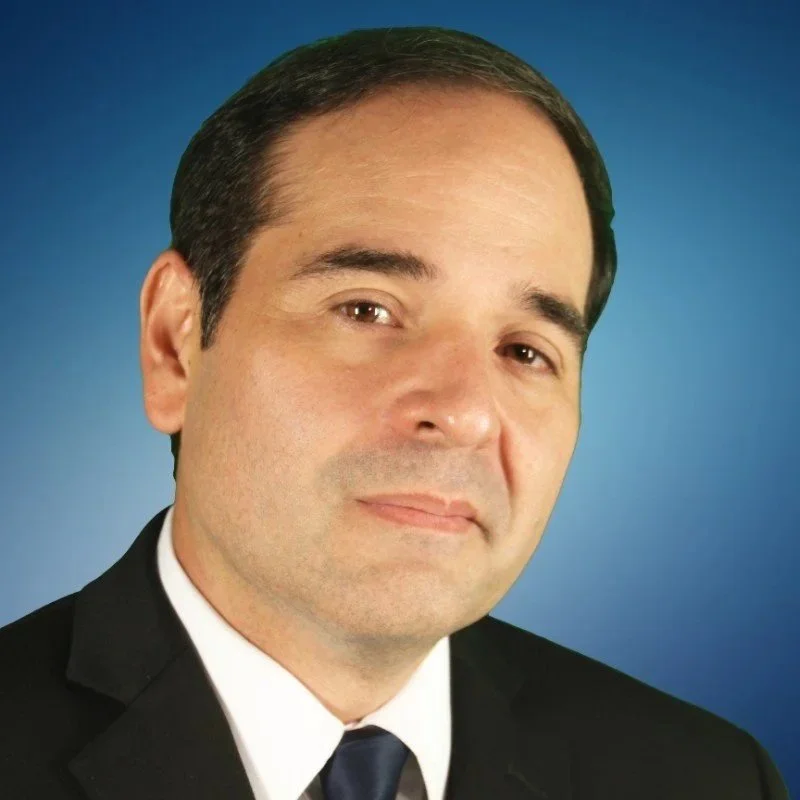 A man wearing a black suit, white shirt, and dark tie against a blue gradient background.