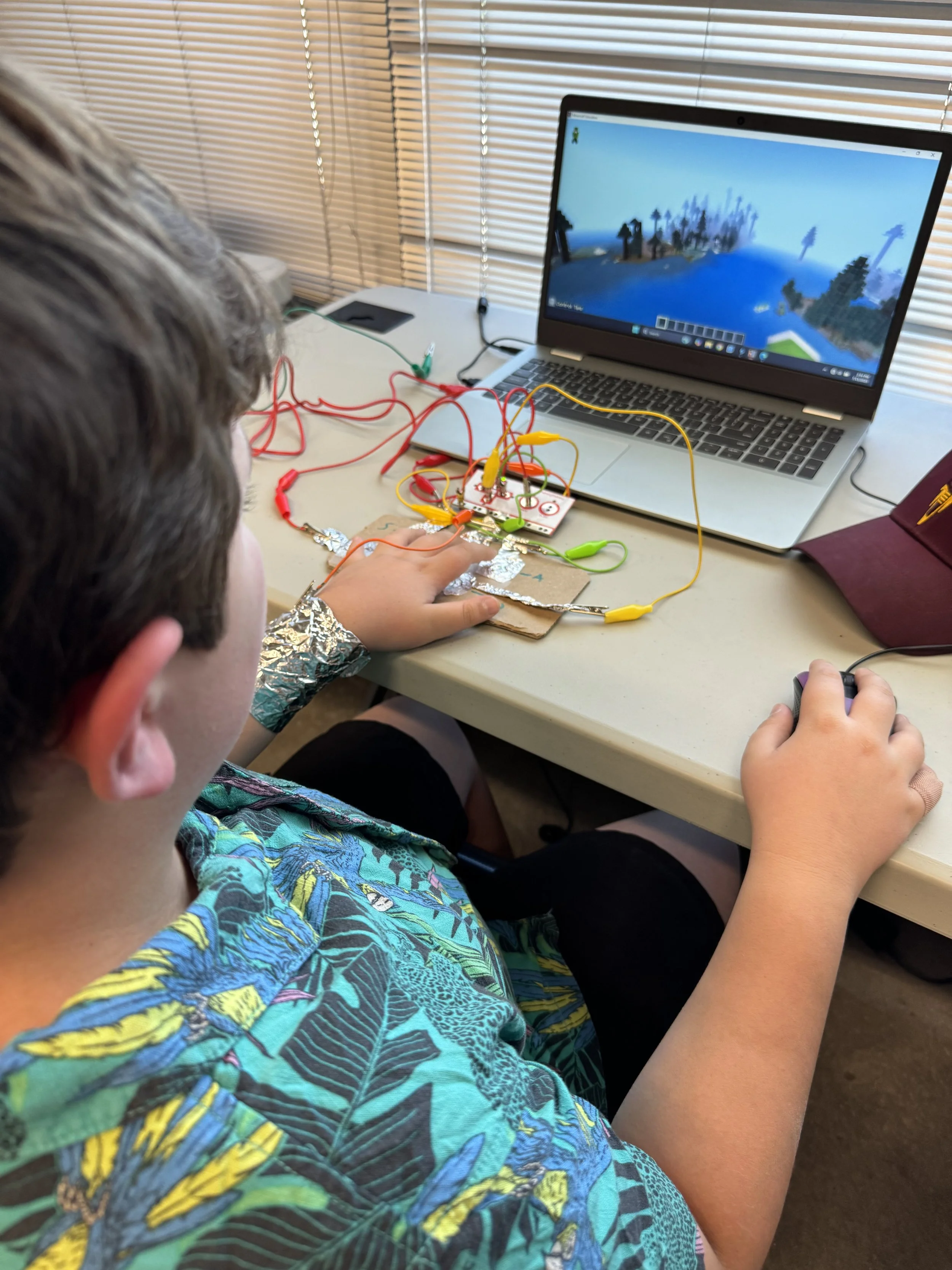 A boy with an aluminum foil wristband is working on an electronics project connected to a laptop displaying a blocky landscape game.