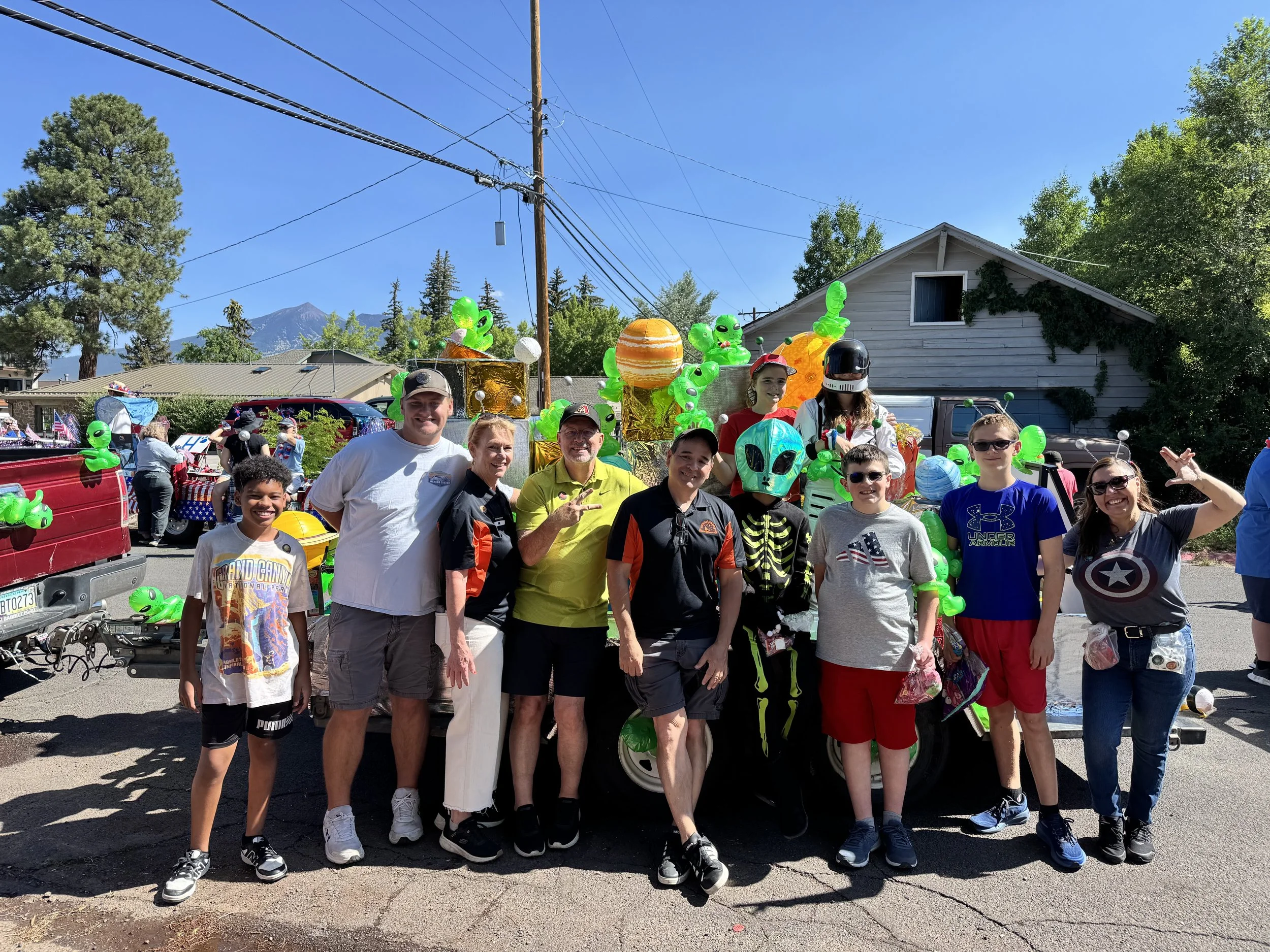A group of people, including children and adults, standing in front of a float decorated with green alien balloons, planets, and space-themed decorations during a parade on a sunny day.