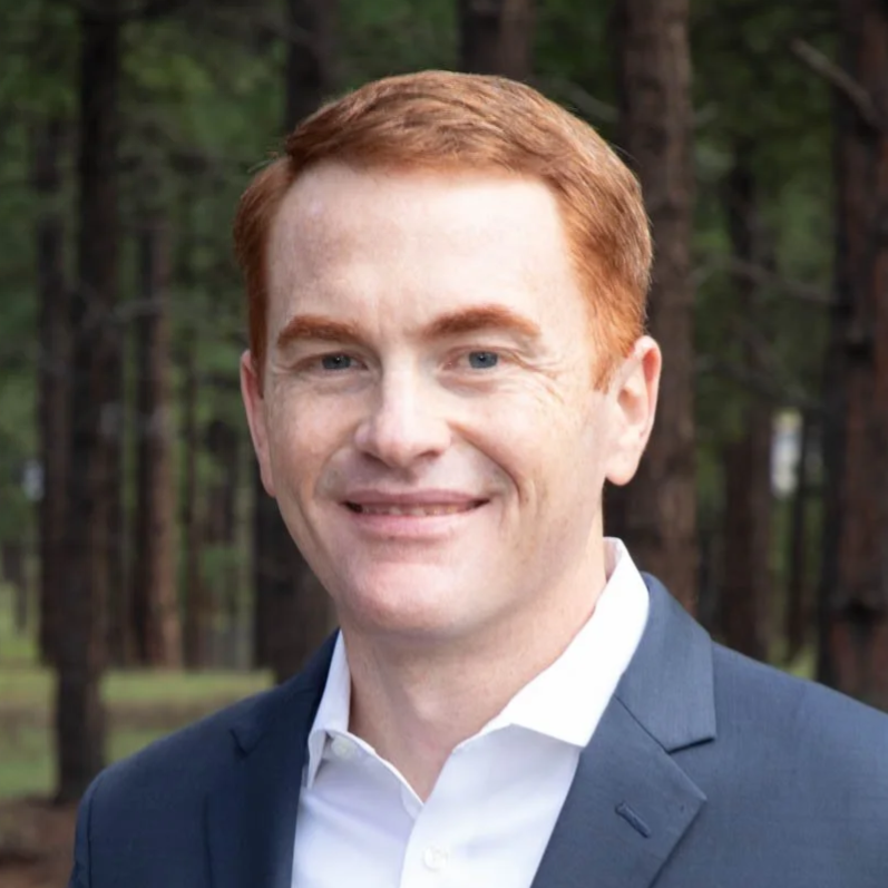 A man with short red hair and blue eyes, wearing a dark suit and white dress shirt, smiling with a forest background.