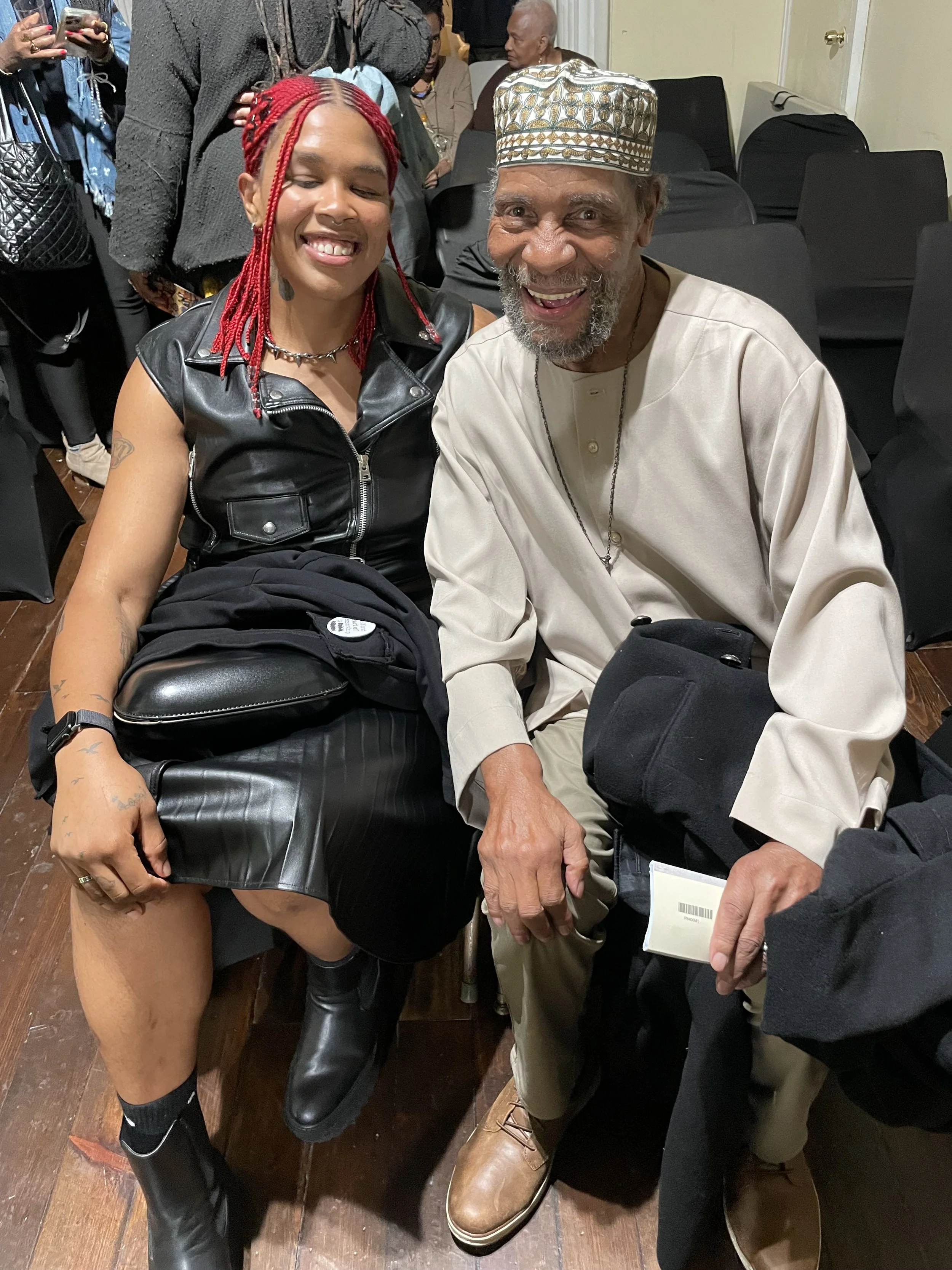 A young woman with red braided hair and a man with a gray beard and traditional cap, both smiling, sitting together at a social gathering.