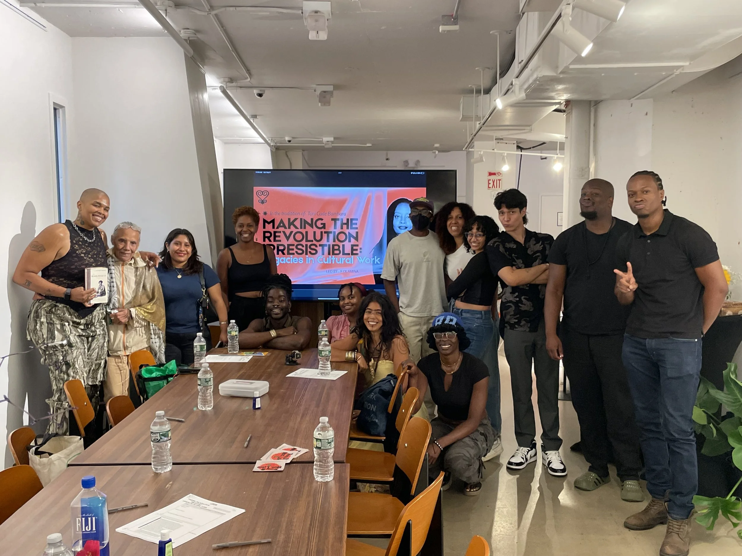 A diverse group of people gathered around a conference table with water bottles, papers, and a camera, smiling for a group photo in front of a large screen displaying a presentation titled 'Making the Revolution Irresistible: Legacies in Cultural Work'.