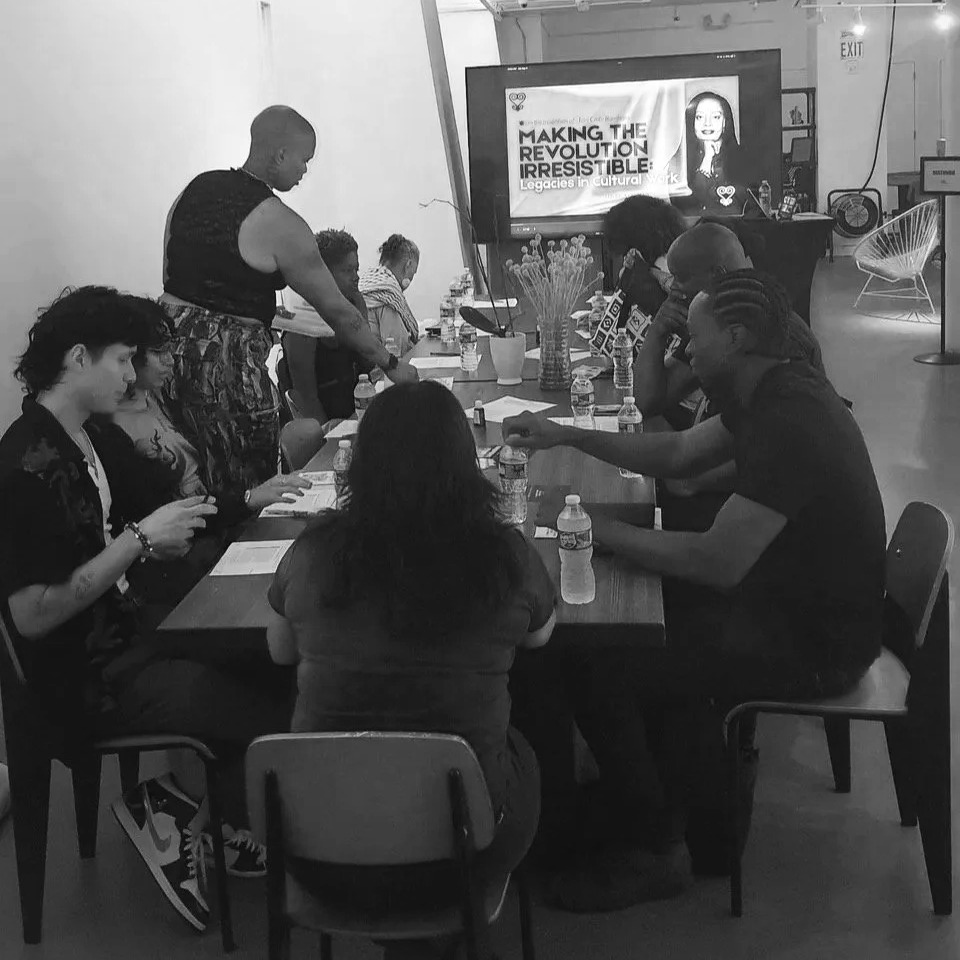 A group of people sitting around a long table with water bottles, in a room with a large screen displaying a presentation titled 'Making the Revolution Irresistible'.